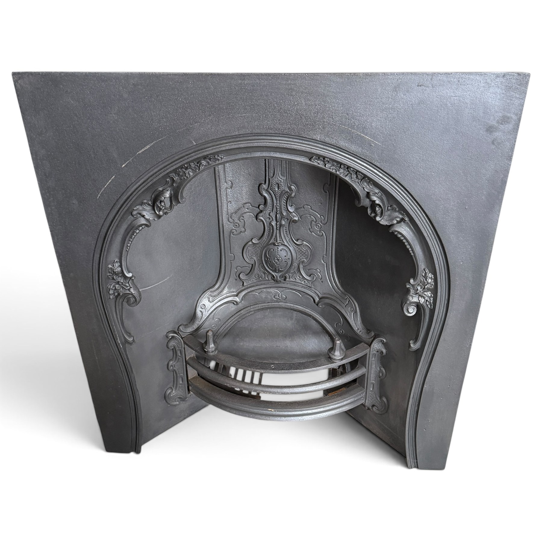 Victorian cast iron fireplace, the arched surround cast with scrolling foliate ornament and central shield-shaped cartouche, fitted with a bowed fire basket, in black painted finish