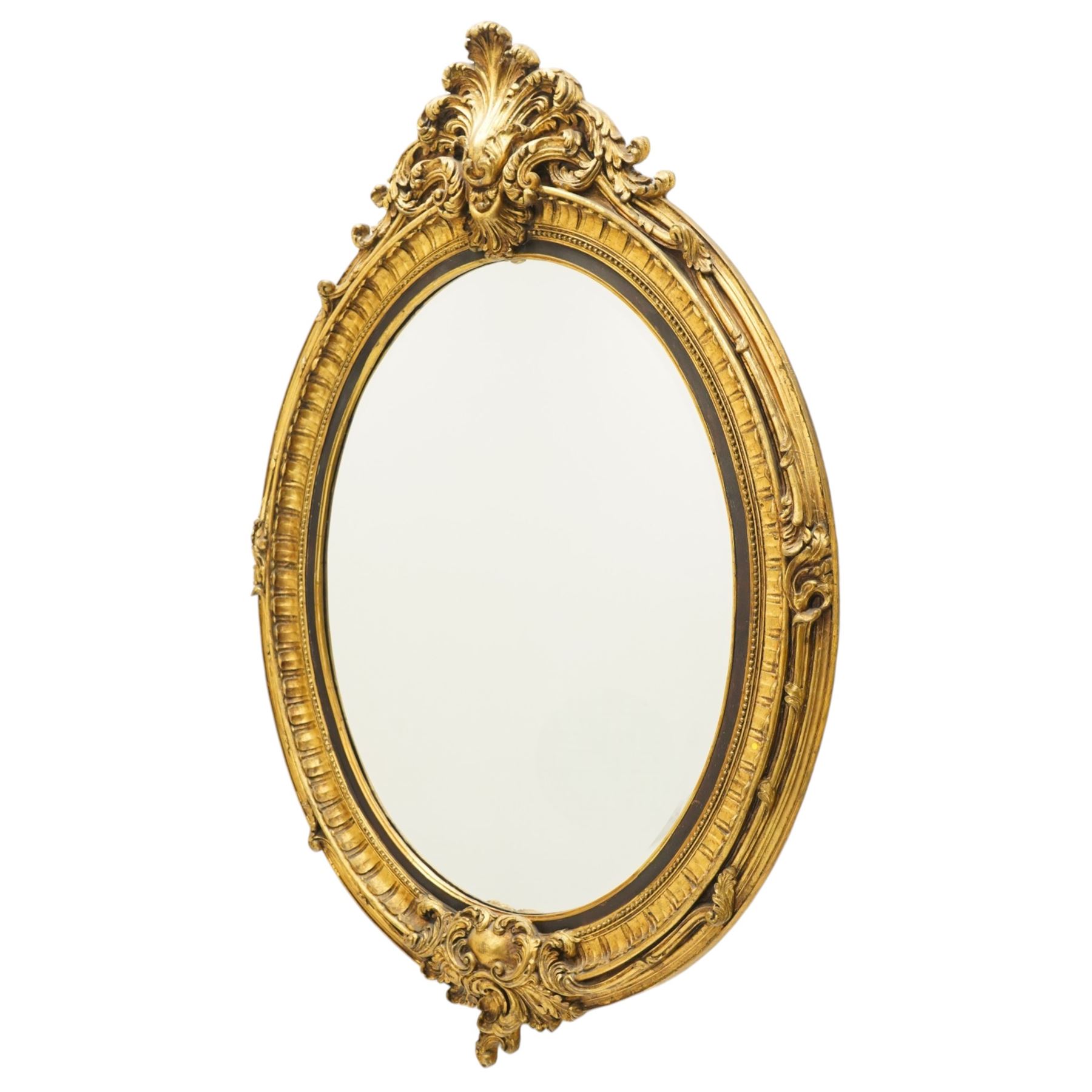 Pair of Rococo Revival gilt framed oval wall mirrors, the exuberant pediment formed of scrolling acanthus leaves and rocaille ornament, the oval bevelled plate set within with ebonised slip with outer repeating lappet decoration, over a rocaille cartouche terminal