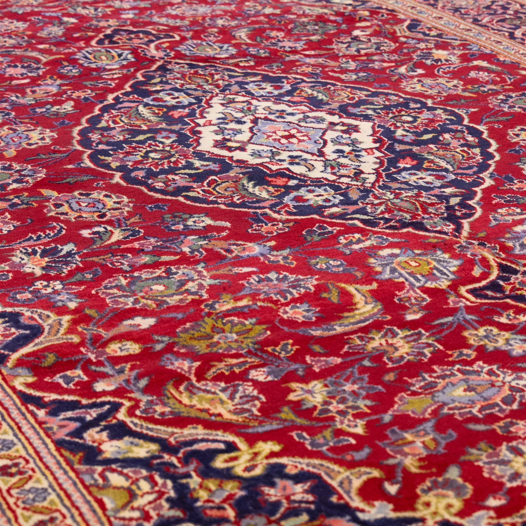Central Persian Kashan hand-knotted rug, crimson red field with central ivory and indigo floral medallion surrounded by an all-over arrangement of finely drawn palmettes, flowering vines, and rosettes, with corresponding navy blue spandrels enclosed by a floral main border with multiple guard stripes, with cotton fringe ends    