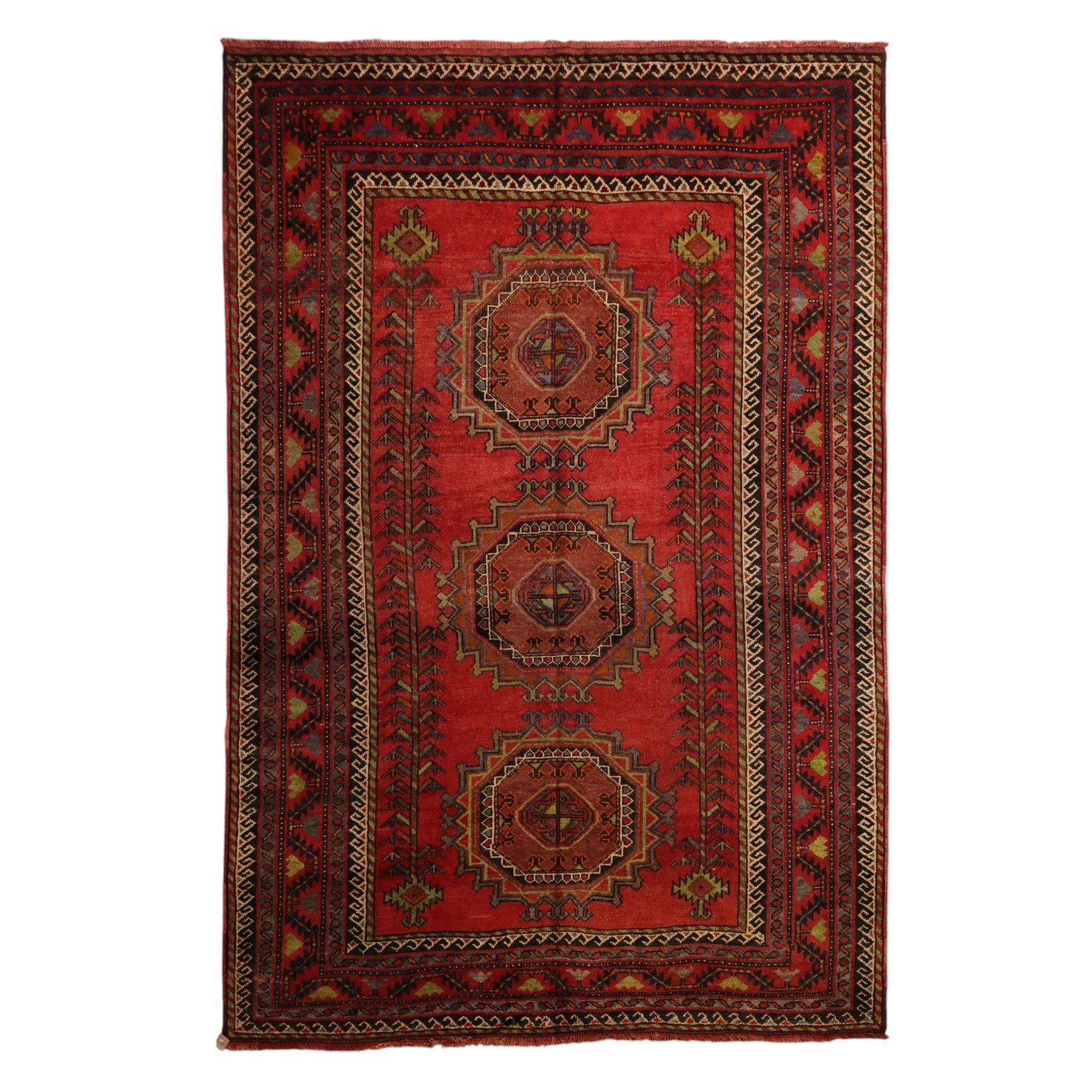 North East Persian Kurdish hand-knotted carpet, madder red field with three vertical and central stepped octagonal medallions enclosing angular geometric motifs with scattered tribal devices, framed by multiple narrow borders with latch-hook patterns, enclosing a bold main border of repeating geometric motifs, palette with indigo, dark brown and soft green tones, 