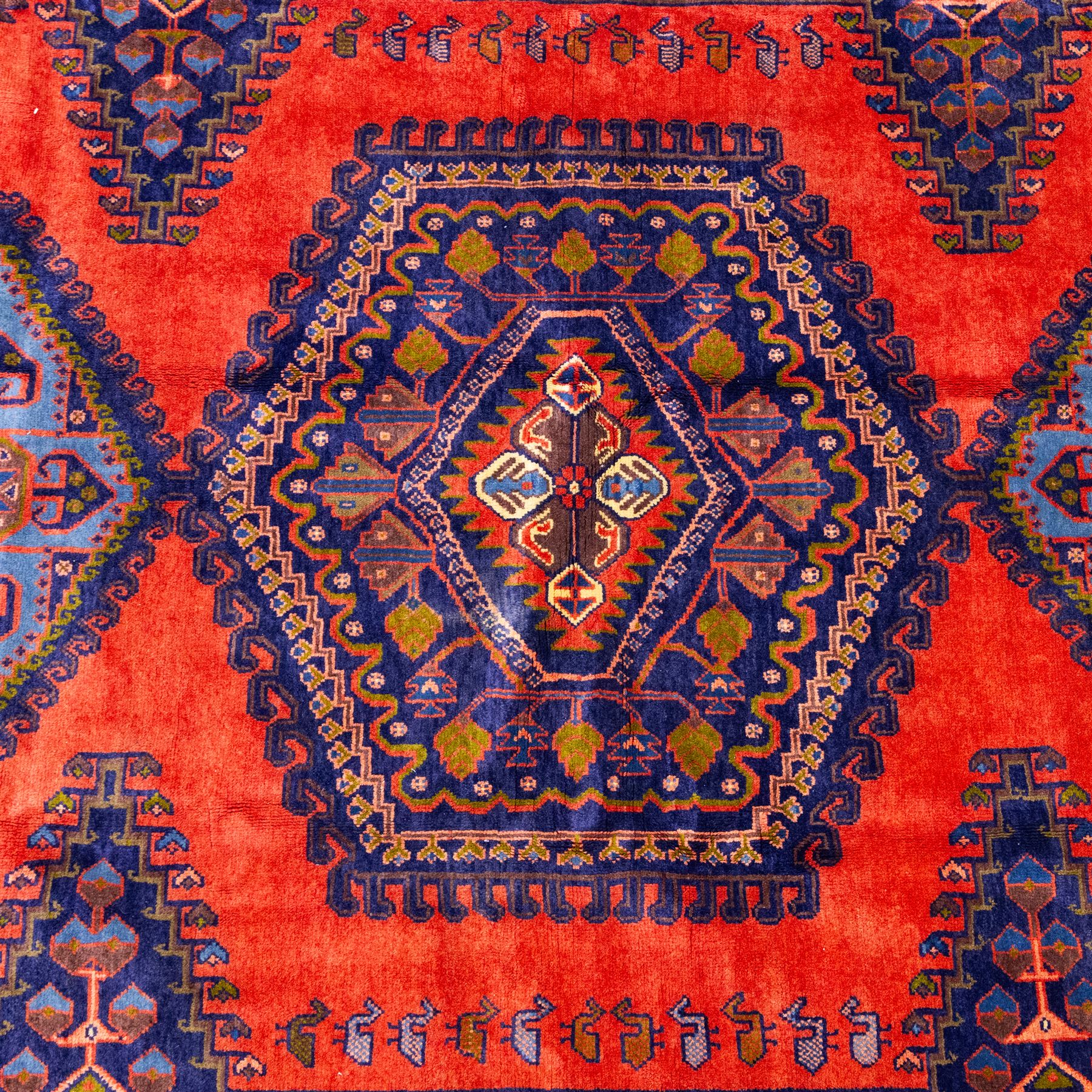 West Persian Veis hand-knotted rug, coral red field decorated with a series of bold geometric lozenge medallions in shades of indigo and sky blue surrounded by angular tribal motifs and stylized animal and plant forms framed by a deep indigo border of geometric ornament and multiple guard stripes, cotton fringe ends 