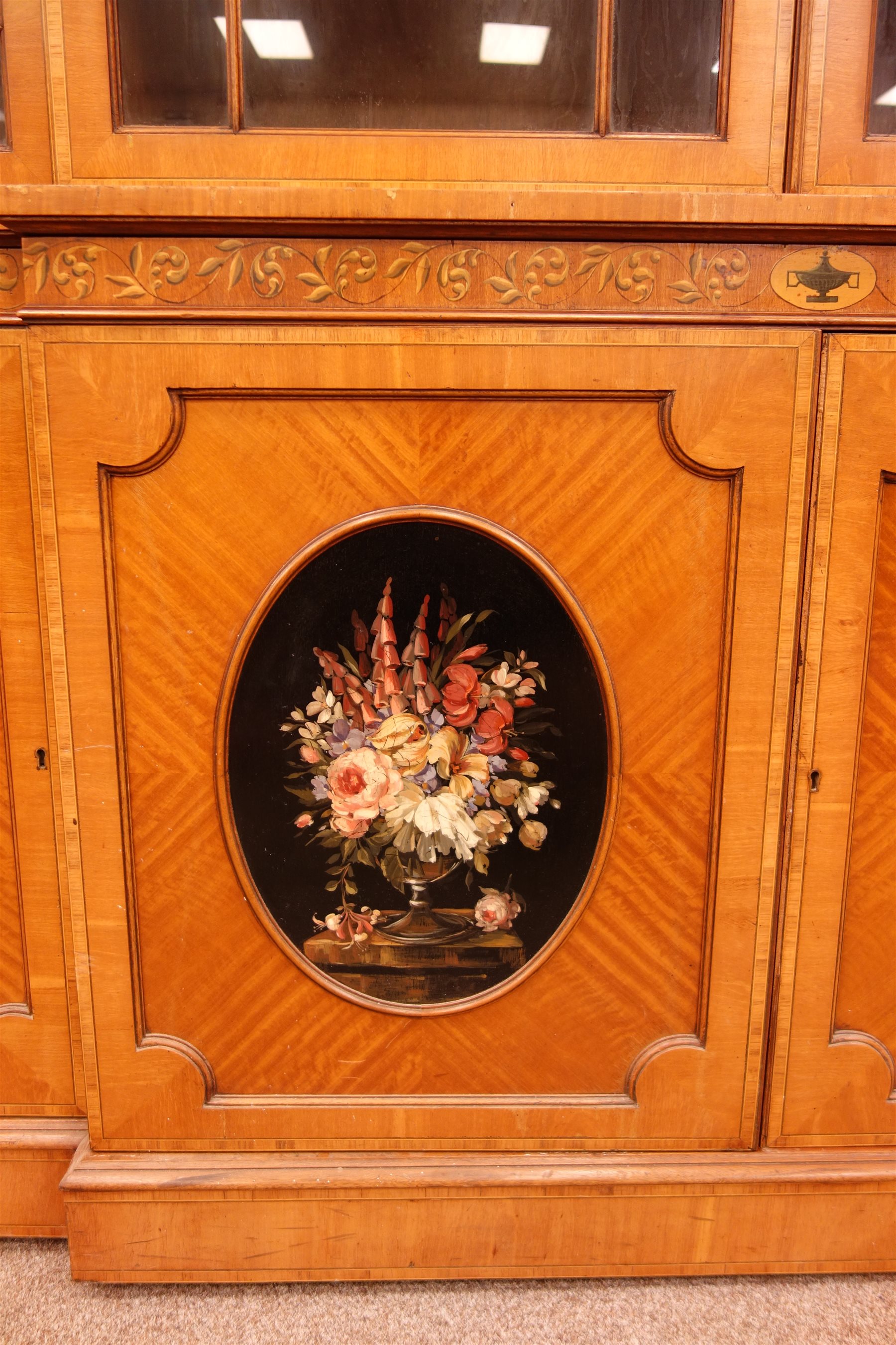 Sheraton Revival satinwood breakfront bookcase, frieze painted with scrolling leafage above four astragal doors with adjustable shelves, the base with four doors painted with floral sprays in oval panels, on a plinth base, W234cm, H235cm, D52cm