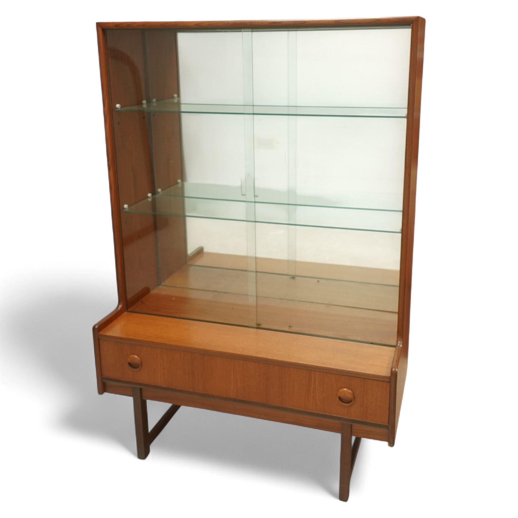 Turnidge of London mid-century teak display cabinet, rectangular top with a pair of glass sliding doors enclosing two adjustable shelves with mirrored back, projecting lower section incorporating one full-width drawer with recessed circular handles, raised on square-section legs with lateral stretchers  