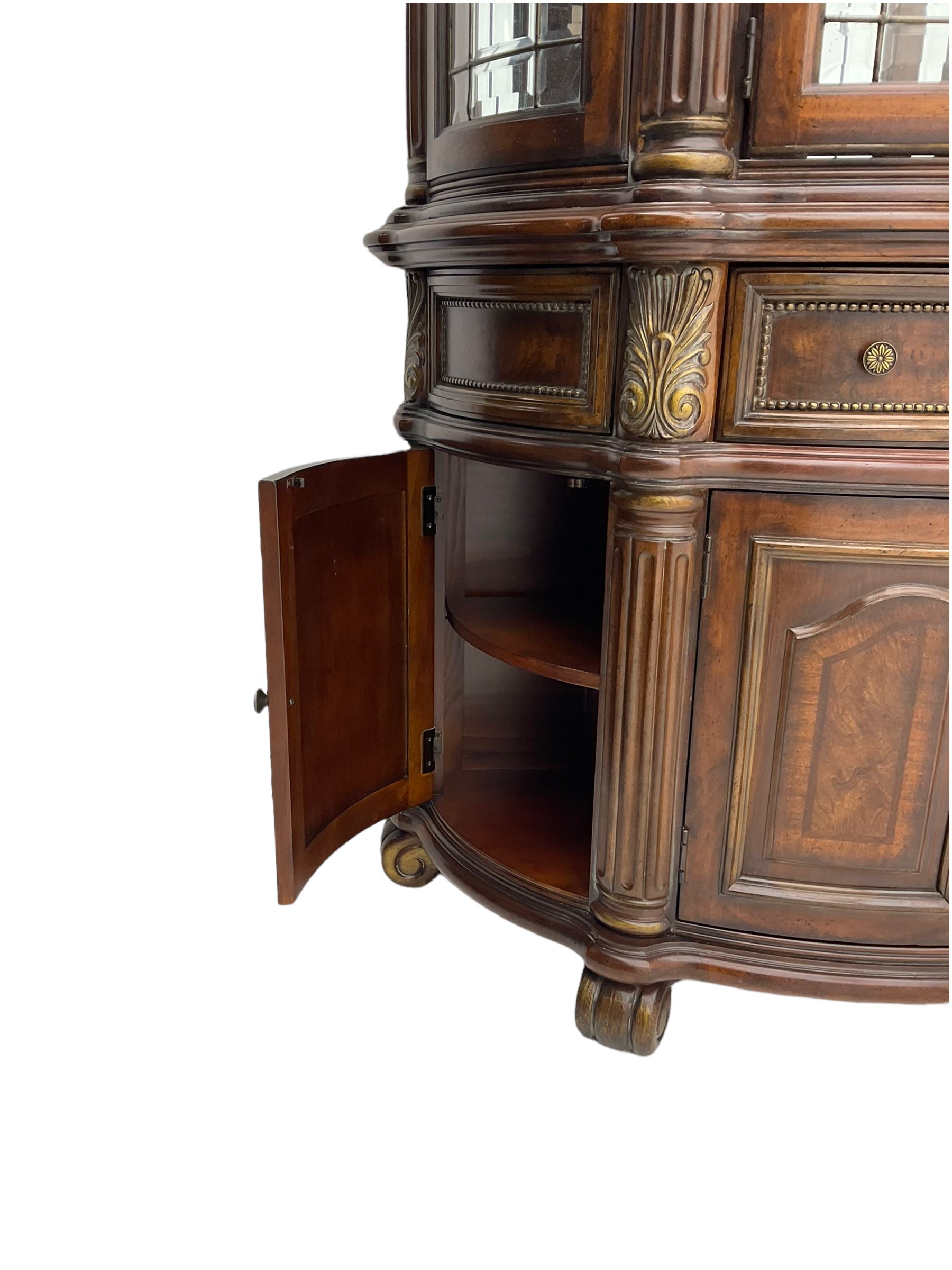  Kevin Charles American walnut demi lune display cabinet with illuminated interior, projecting cornice, two bevel edge glazed doors enclosing two adjustable shelves above single drawer and four cupboard doors with wine rack, carved scrolling supports