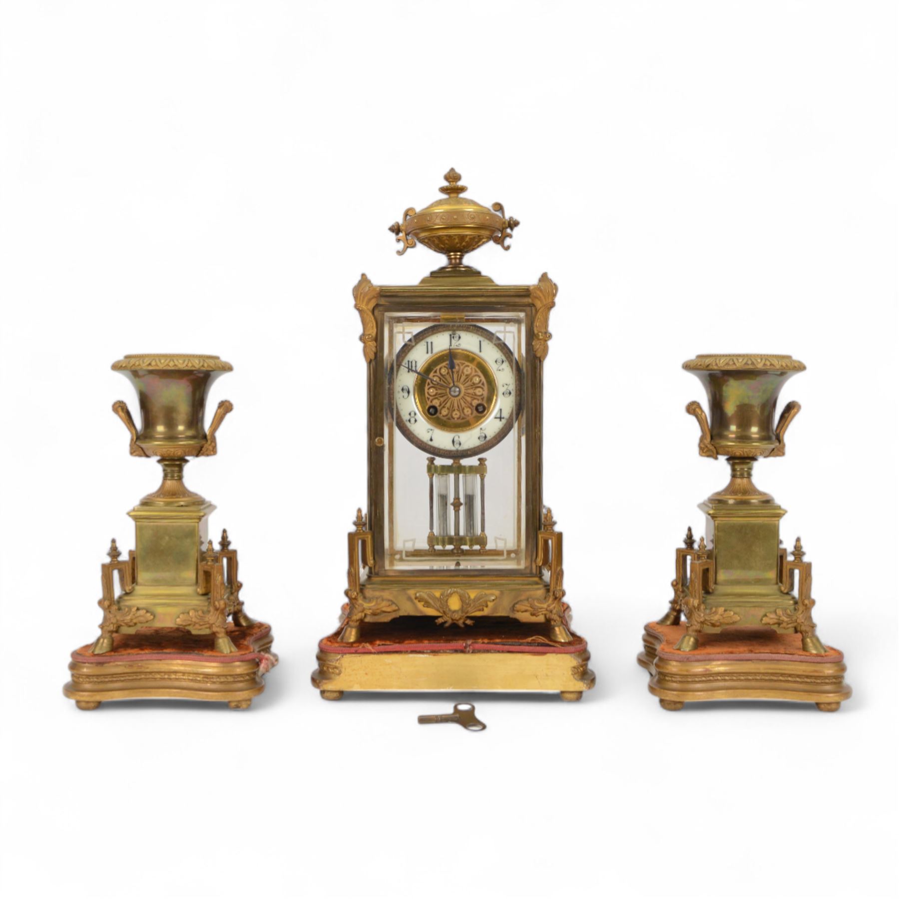 French- Art-deco Edwardian 8-day four-glass clock garniture, with etched glass panels, cast brass decoration to the corners of the case and splayed feet representing cloven hoofs to the base, case surmounted by a large oval urn on a tiered brass platform, two part dial with a recessed brass centre comprising of pierced radial decoration, with steel spade hands and Arabic numerals to the chapter, rack striking movement with a deadbeat Brocot escapement, striking the hours and half-hours on a bell. With a visible twin file mercury pendulum. Garni comprising two large twin handled urns on rectangular pedestals raised on conforming feet representing cloven hoofs, Clock and garni presented on three shaped wooden gesso bases with velvet pads. With key.
