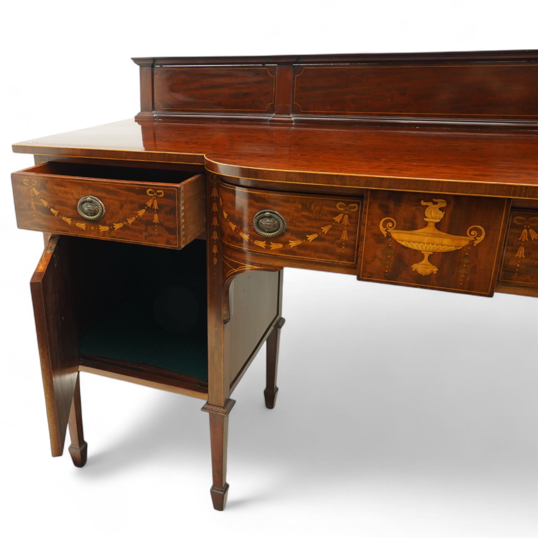 Edwardian Sheraton Revival inlaid plum pudding mahogany serpentine sideboard, rectangular top with raised gallery above bowed frieze drawer finely inlaid with central neoclassical urn motif, flanked by two drawers with swags and ribbon bows, above a cellarette drawer and cupboard, on square tapered supports with spade feet 