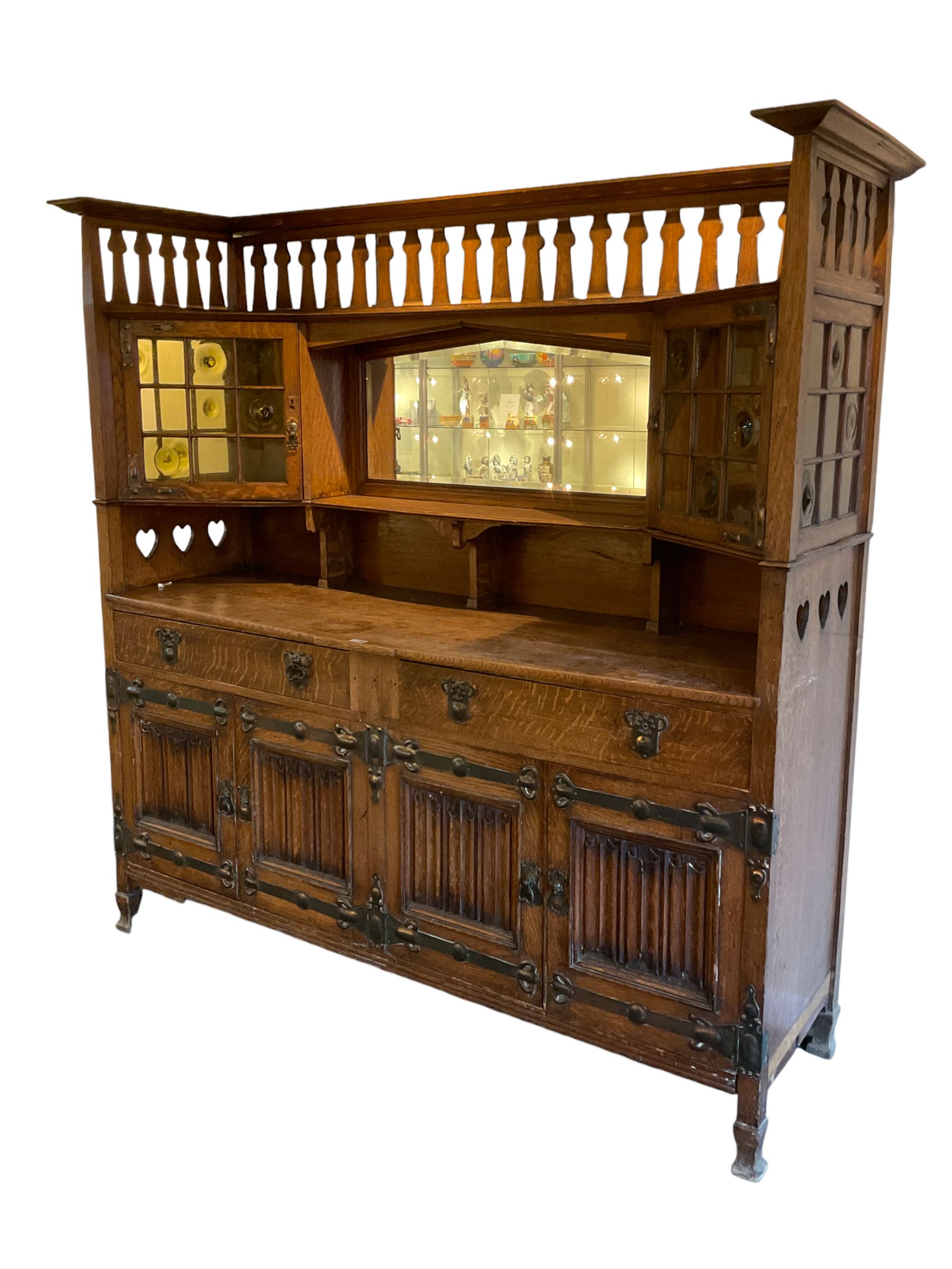Shapland and Petter - Arts & Crafts period oak dresser, raised gallery top with spade balustrade, central bevelled mirror flanked by two cabinet, astragal glazed doors with yellow tinted glass and bullseye panes, the sides with pierced heart shaped decoration, fitted with two drawers and two double cupboards, enclosed by linenfold panelled doors,  copper work strap hinges and fixtures, on shaped spade feet, the locks stamped 'S&P'