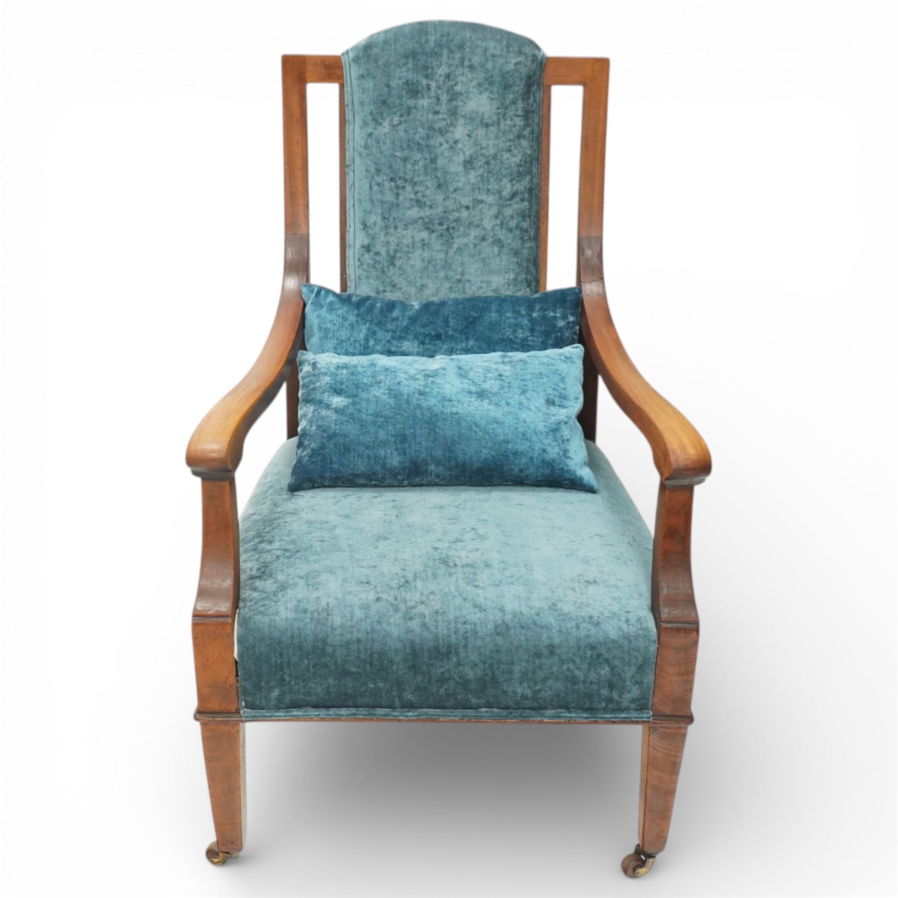 Arts & Crafts mahogany framed armchair, shaped rectangular back with open uprights, padded back, seat and arm supports upholstered in blue velvet, downswept arms on square tapering front supports with brass caps and castors, the rear supports slightly splayed