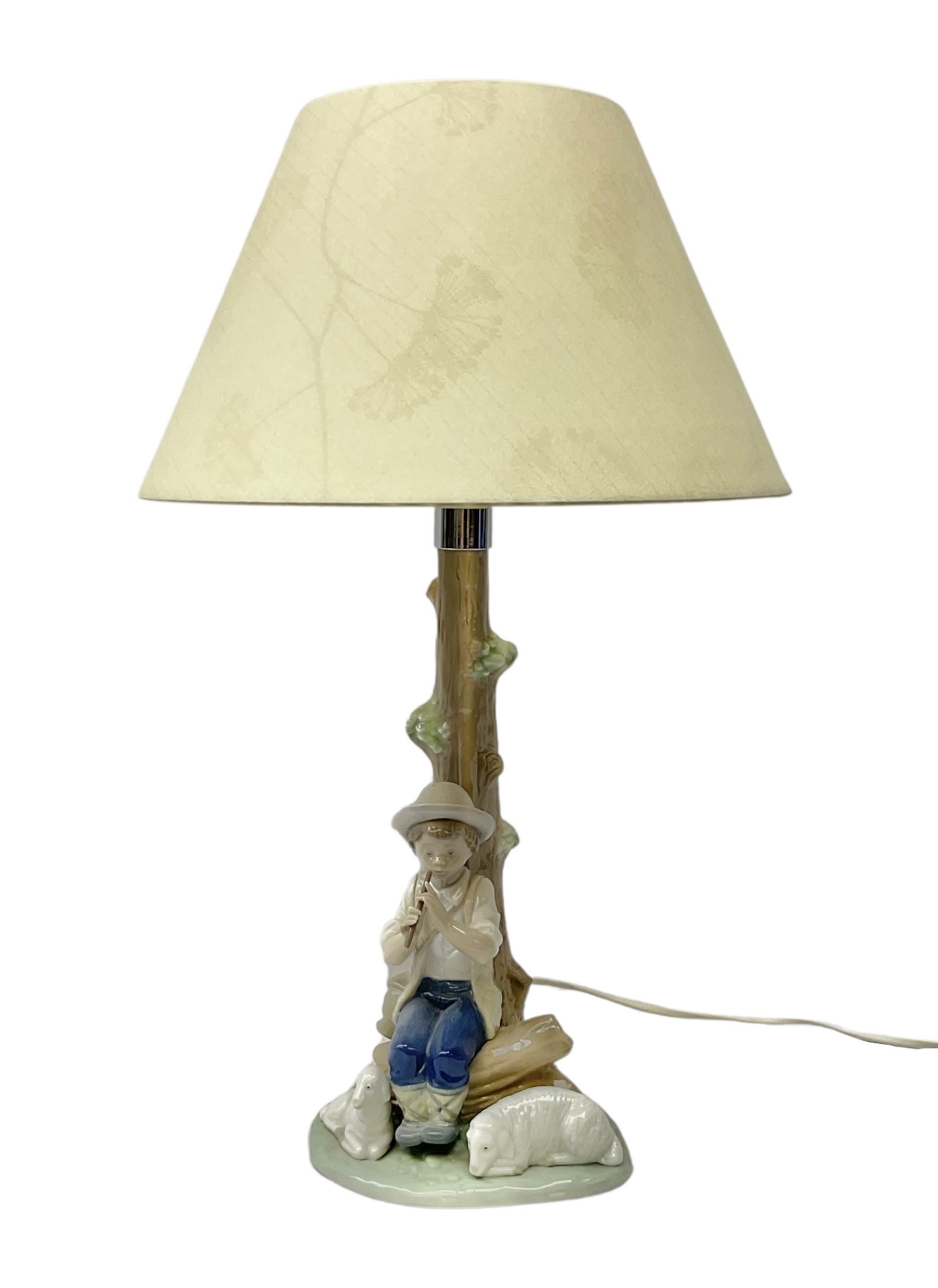 Nao table lamp modelled as a boy playing the flute upon logs with two recumbent sheep at his feet, the stem modelled as a tree trunk, with fabric shade, with printed mark beneath, H46cm