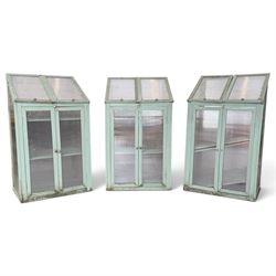 Three green-painted timber cold-frame greenhouses or growhouses, each with twin hinged sloping polycarbonate lids above a pair of panelled doors enclosing slatted shelves