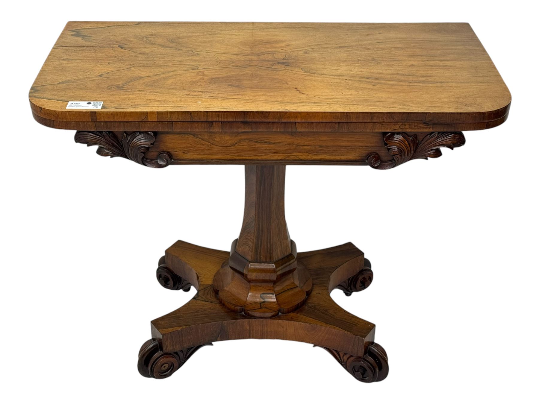 Early Victorian rosewood card table, rectangular foldover swivel top with rounded corners, red baize-lined surface, frieze adorned with carved foliate motifs, supported by a central octagonal waisted column on shaped quatrefoil base, terminating in scrolled feet with concealed metal castors