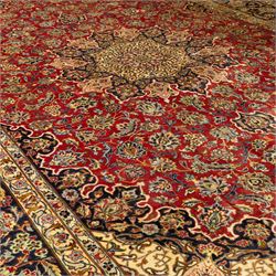 Central Persian Kashan hand-knotted carpet, crimson ground, with a large central ivory and dark navy floral medallion with pendants, surrounded by dense scrolling foliate and floral motifs incorporating rosettes, palmettes and vine work, enclosed by ivory spandrels and a dark navy border with repeating palmettes and meandering vines, flanked by floral guard stripes