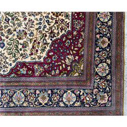 Central Persian Ghom hand-knotted rug, ivory field with scrolling vines, palmettes and flowerheads around a central lobbed medallion with pendants, framed by burgundy spandrels and a navy border of rosettes and arabesques, 289cm x 214cm