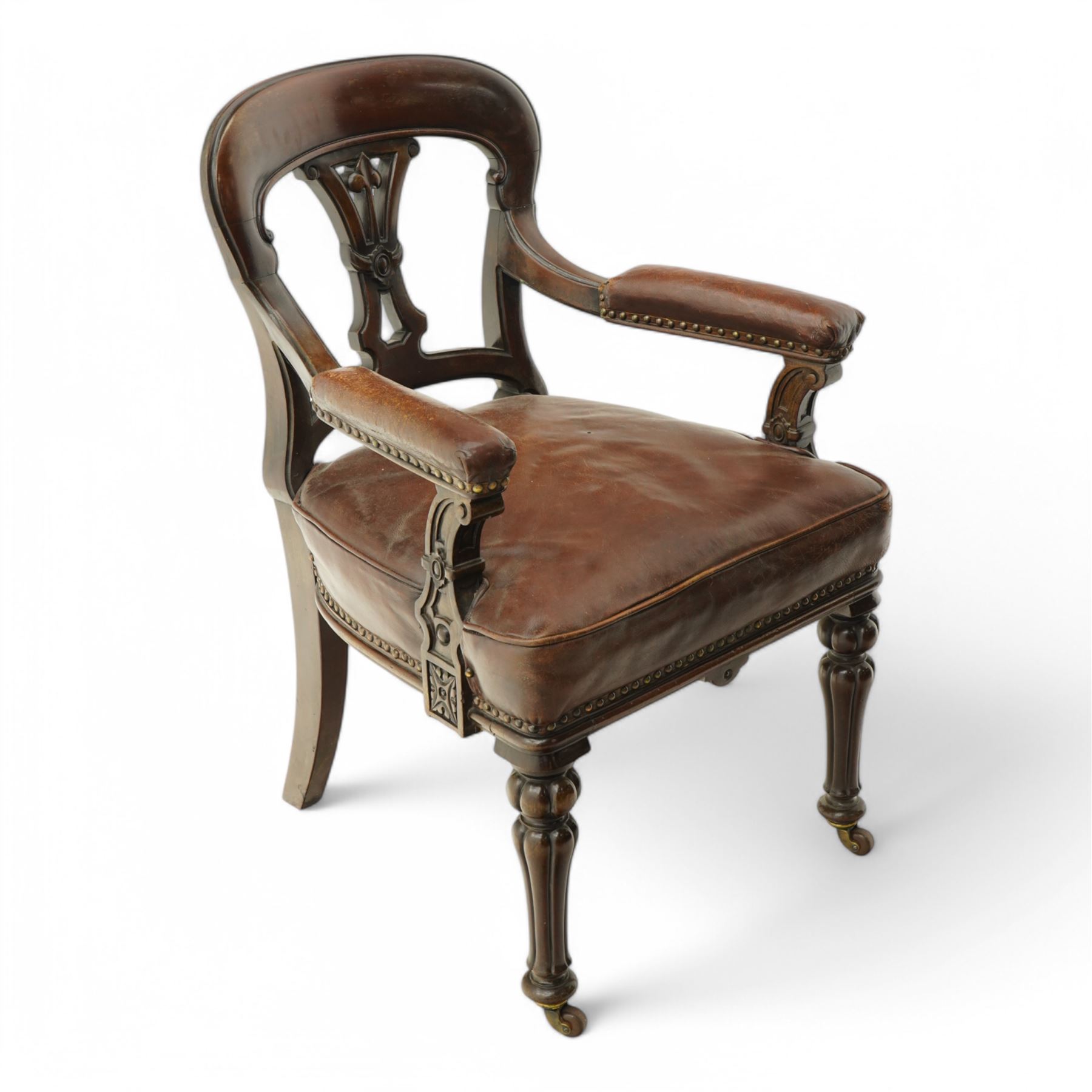 Victorian mahogany library elbow chair, arched cresting rail over shaped and pierced splat carved with cartouche, upholstered arms and seat in brown leather with studded bands, turned and lobe carved front supports on brass and ceramic castors 