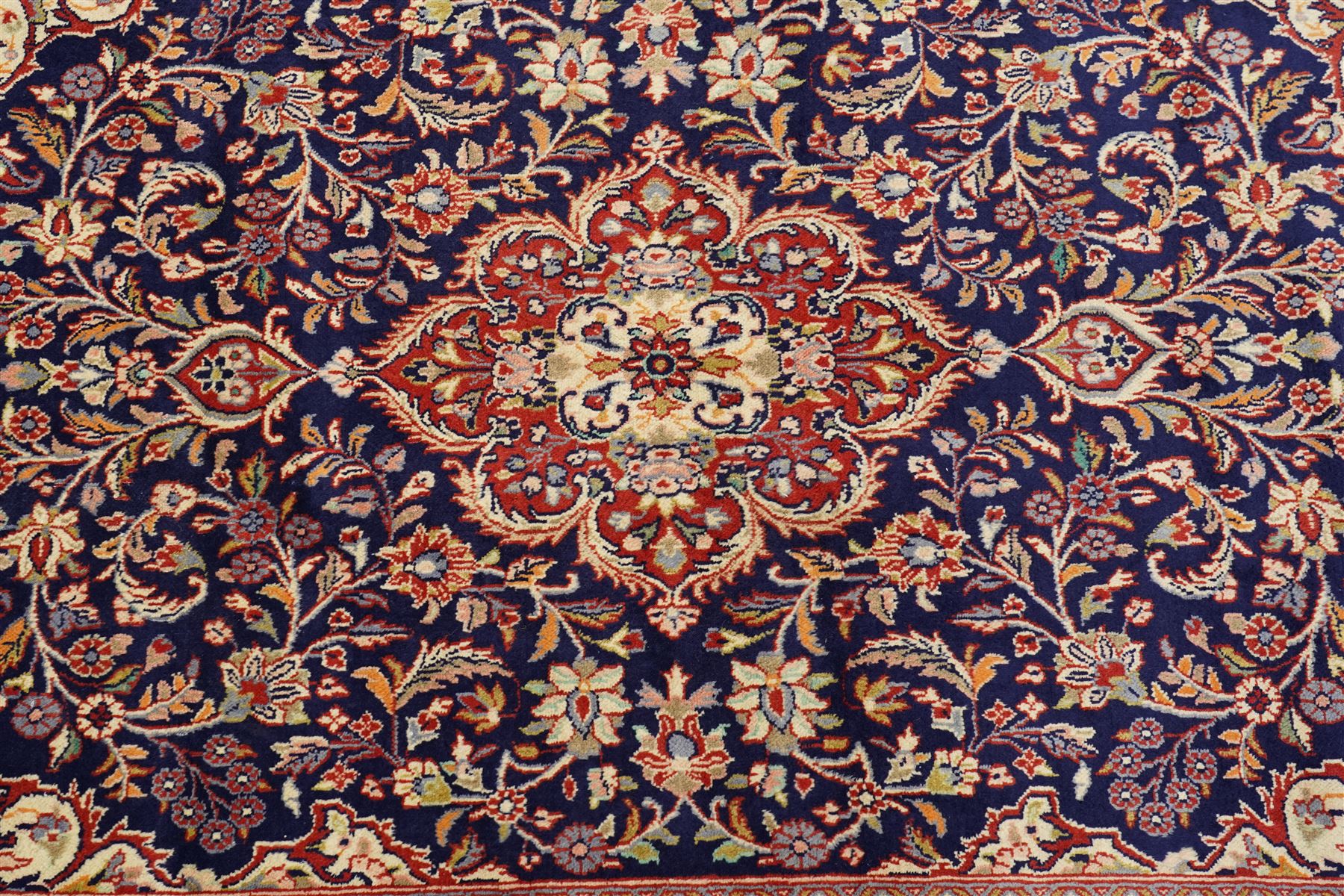 Central Persian Kashan hand-knotted rug, indigo field with central lobed medallion with pendants issuing scrolling arabesque of palmettes, flowering vines, and foliate tendrils, with conforming spandrels, with vine border and framed by guard stripes in tones indigo, ivory and madder red, with cotton fringe ends 