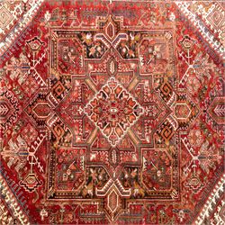 North West Persian Heriz hand-knotted carpet, madder red field, with a large angular stepped central medallion with serrated outlines, flanked by bold stylized palmettes, hooked leaves and geometric devices, spandrels similarly decorated within a wide geometric main border featuring angular floral motifs, enclosed by multiple guard borders in madder red, indigo blue, ivory, olive green and soft ochre, with cotton fringe ends 