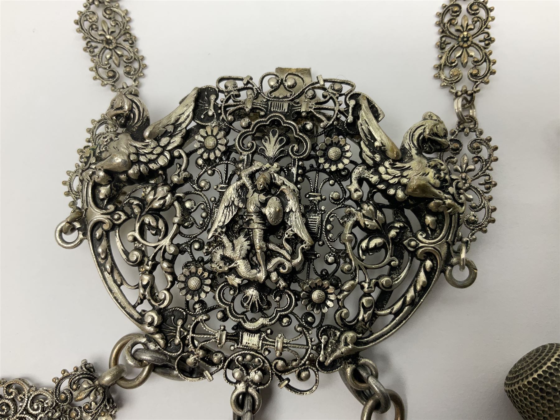 19th century continental silver plated chatelaine, the main body flanked with winged mythical beasts and central soldier slaying a dragon supporting three suspended items to include velvet lined small basket and long clasped filigree chain, together with a thimble