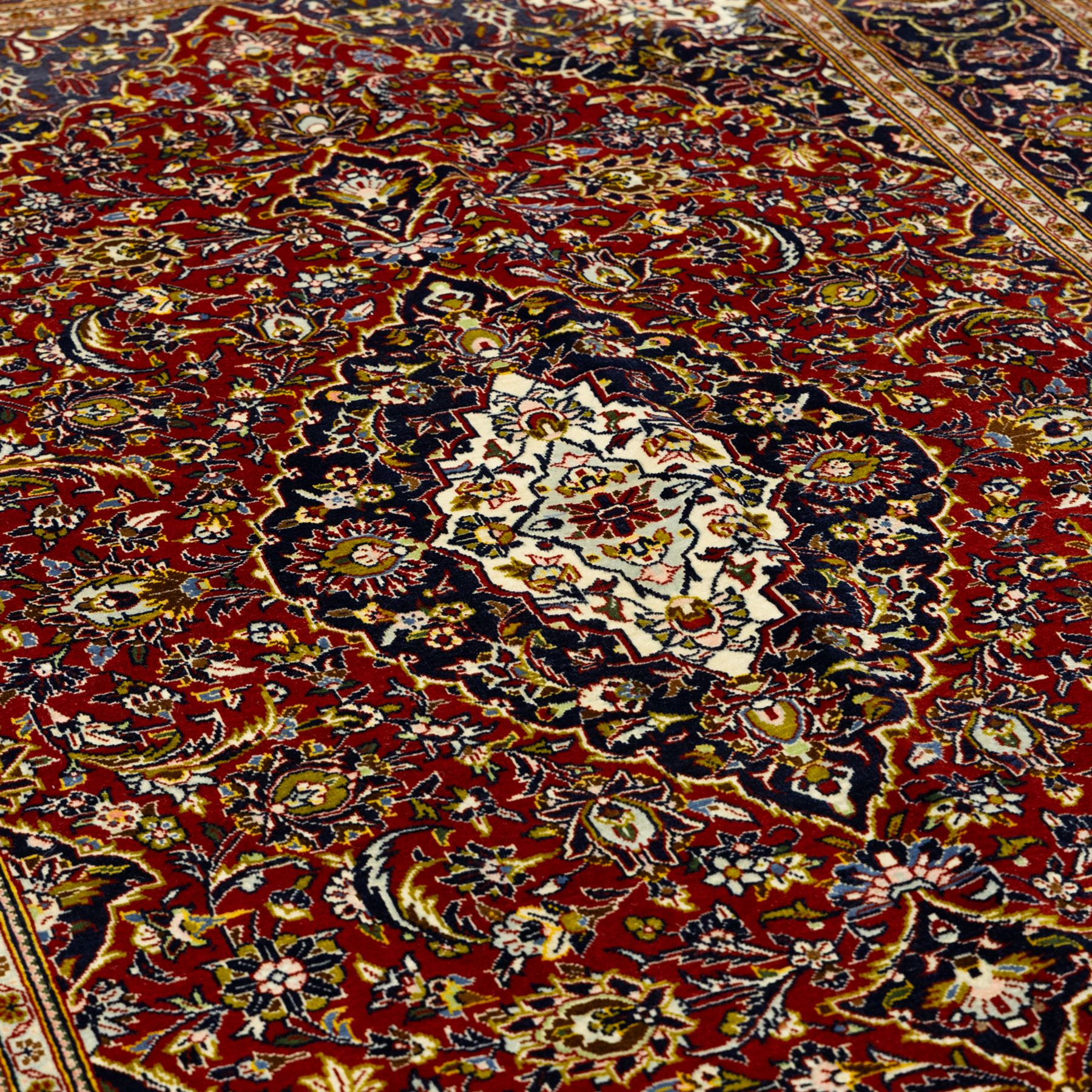 Central Persian Kashan hand-knotted carpet, crimson ground, with a central ivory shaped floral medallion with pendants, surrounded by dense scrolling foliate and floral motifs incorporating rosettes, palmettes and delicate vine work, enclosed by ivory spandrels and a dark navy border with repeating palmettes and meandering vines, flanked by floral guard stripes, with cotton fringes