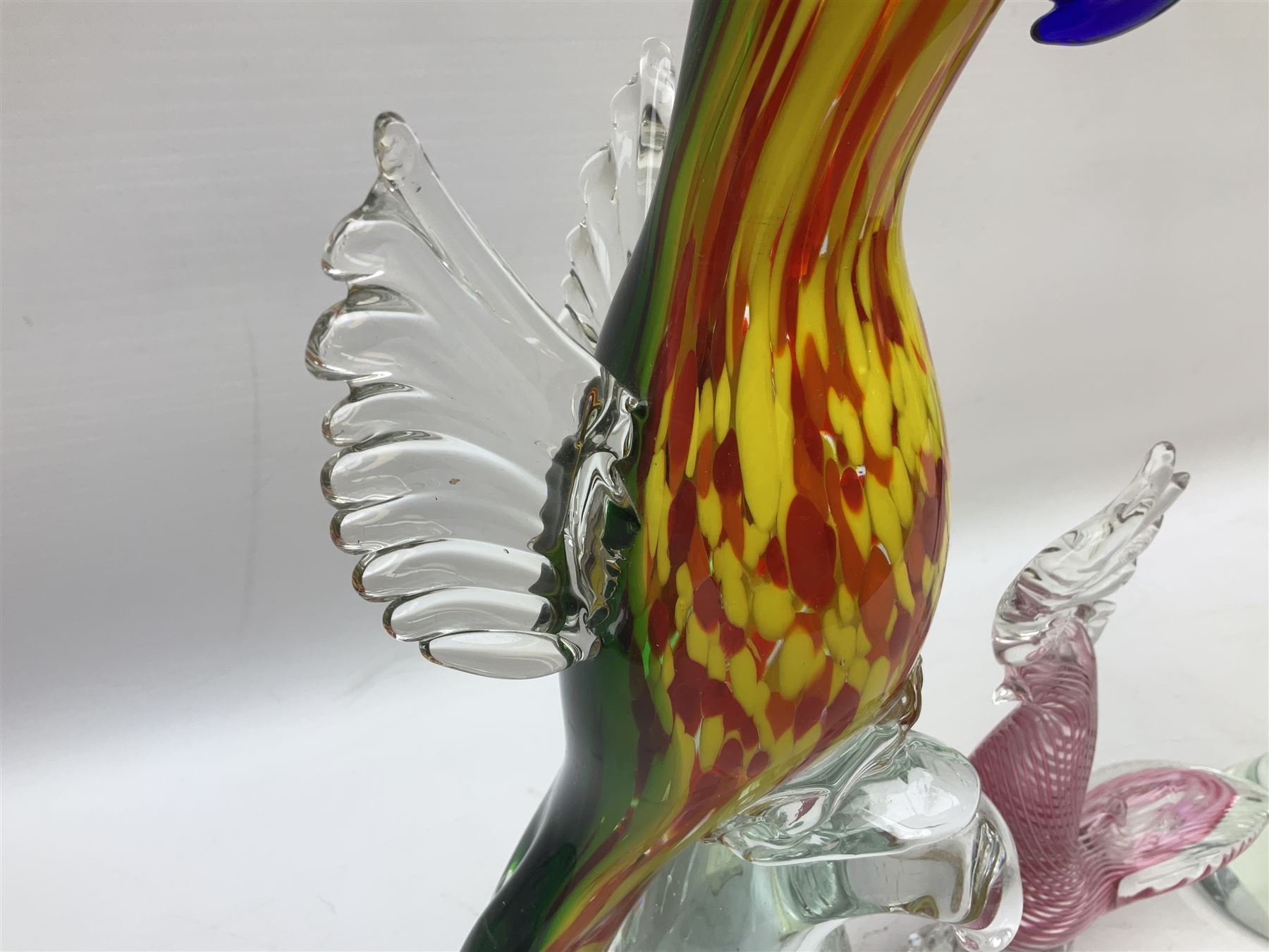 Three Murano style glass cockatoo parrots, the first with mottled yellow, red and orange body, clear outstretched wings, dark blue beak and red crest, raised upon clear stylised base, the second smaller parrot with merging blue and green body, and further with pink striped body, tallest H40cm