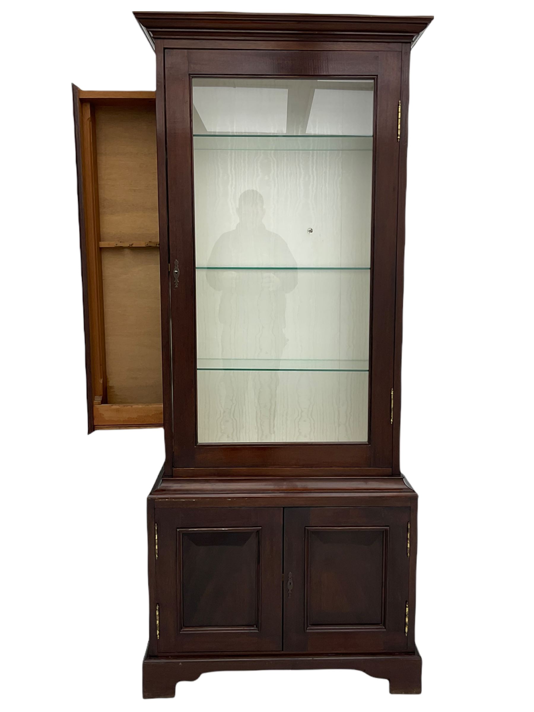 Mahogany display cabinet with hidden compartment, projecting cornice over single glazed door enclosing glass shelves, double panelled cupboard below, the interior with button releasing sliding back compartment, on bracket feet