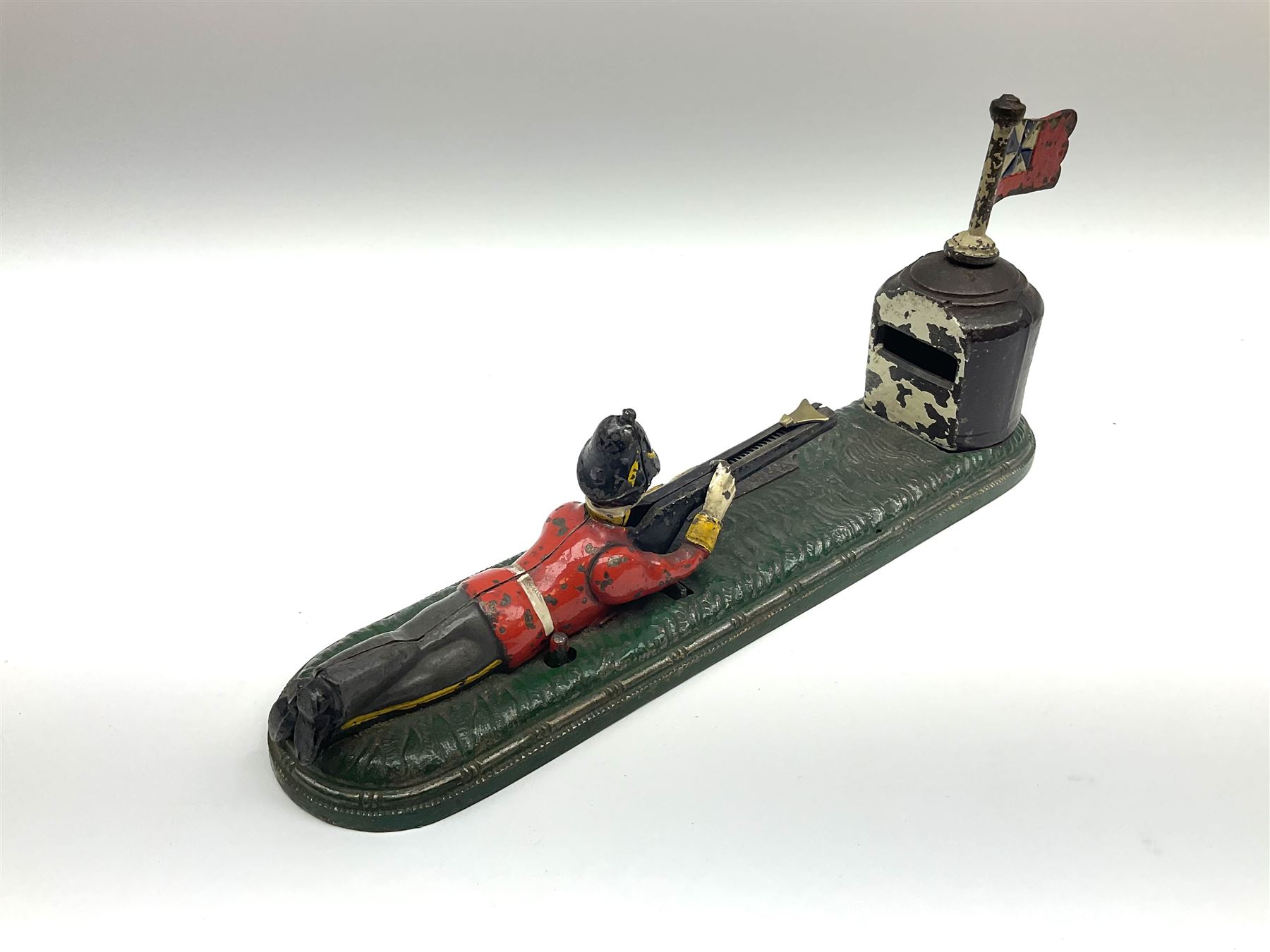 Very rare 19th century John Harper & Co. mechanical cast-iron money box, registered number 33821 patented 1885, inscribed on metal plaque 'Wimbledon Bank', based on the Queen's Trophy for shooting at Wimbledon, depicting a British Infantry man in red tunic and blue trousers lying on the ground using a gun to fire a brass coin launcher into a pill box with removable moving flag above L30cm