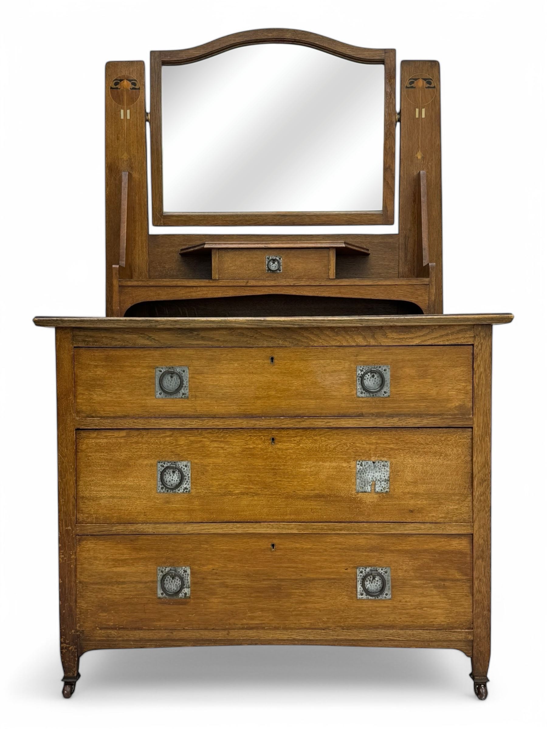 Early 20th century Arts and Crafts oak dressing chest, the arched mirror flanked by floral marquetry inlaid uprights above a shelf and drawer, over three long drawers with square hammered metal plates and circular pulls, on slender supports with castors