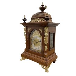 Junghans-German early 20th century walnut cased 8-day mantle clock, with an ogee carved pediment and turned wooden finials, break arch door with brass spandrels beneath flanked by brass caryatids, glazed panels to the sides on a stepped plinth raised on  brass paw feet,  brass dial with a matted centre and silvered chapter with Arabic numerals and Fleur-di Lis steel hands, twin train rack striking movement with a ting tang chime on the quarters. With Key and pendulum. 