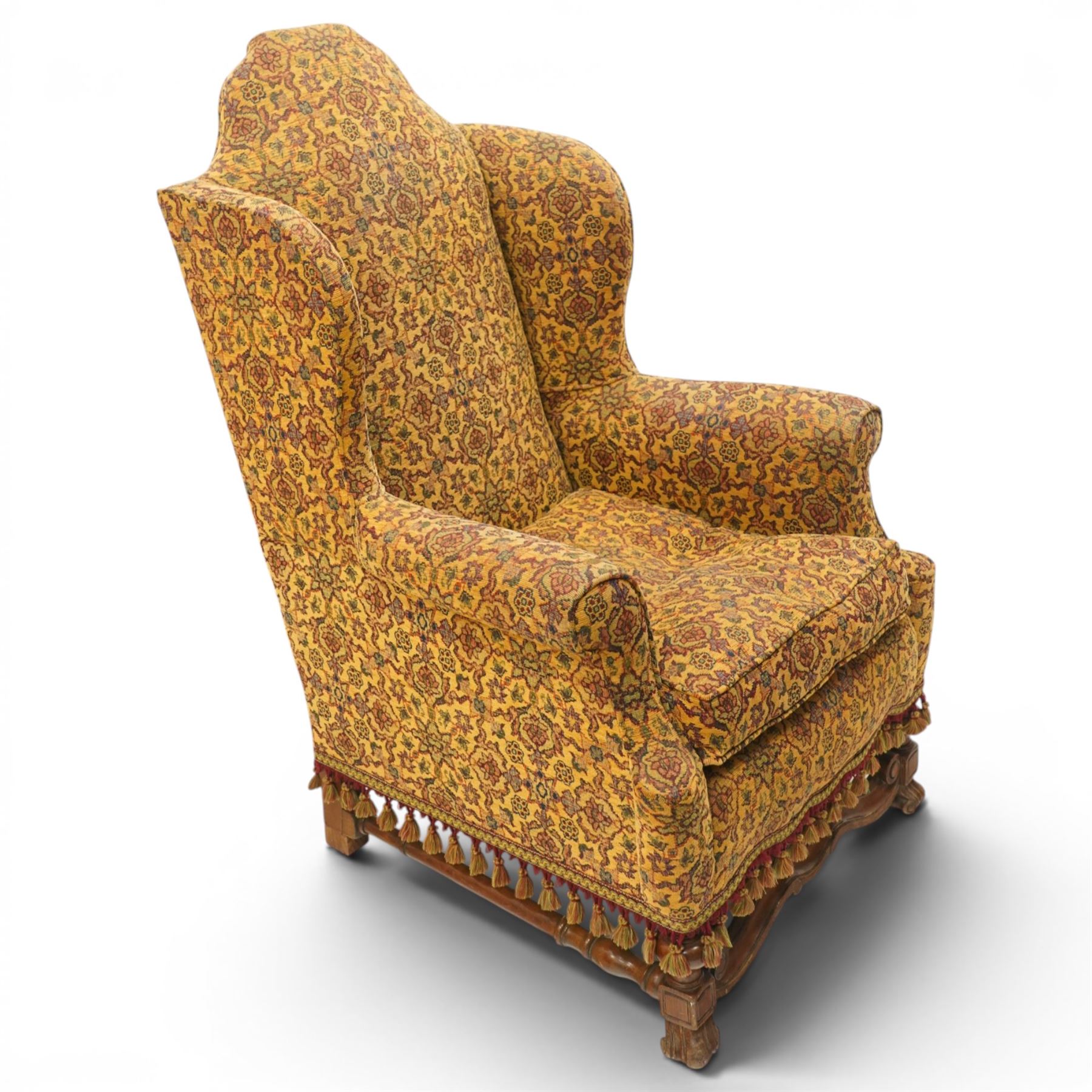 Pair of William & Mary design wingback armchairs, the arched high-back over rolled arms with a loose seat cushion and sprung seat, upholstered in old-gold ground foliate patterned fabric, raise on scroll feet united by a front serpentine stretcher