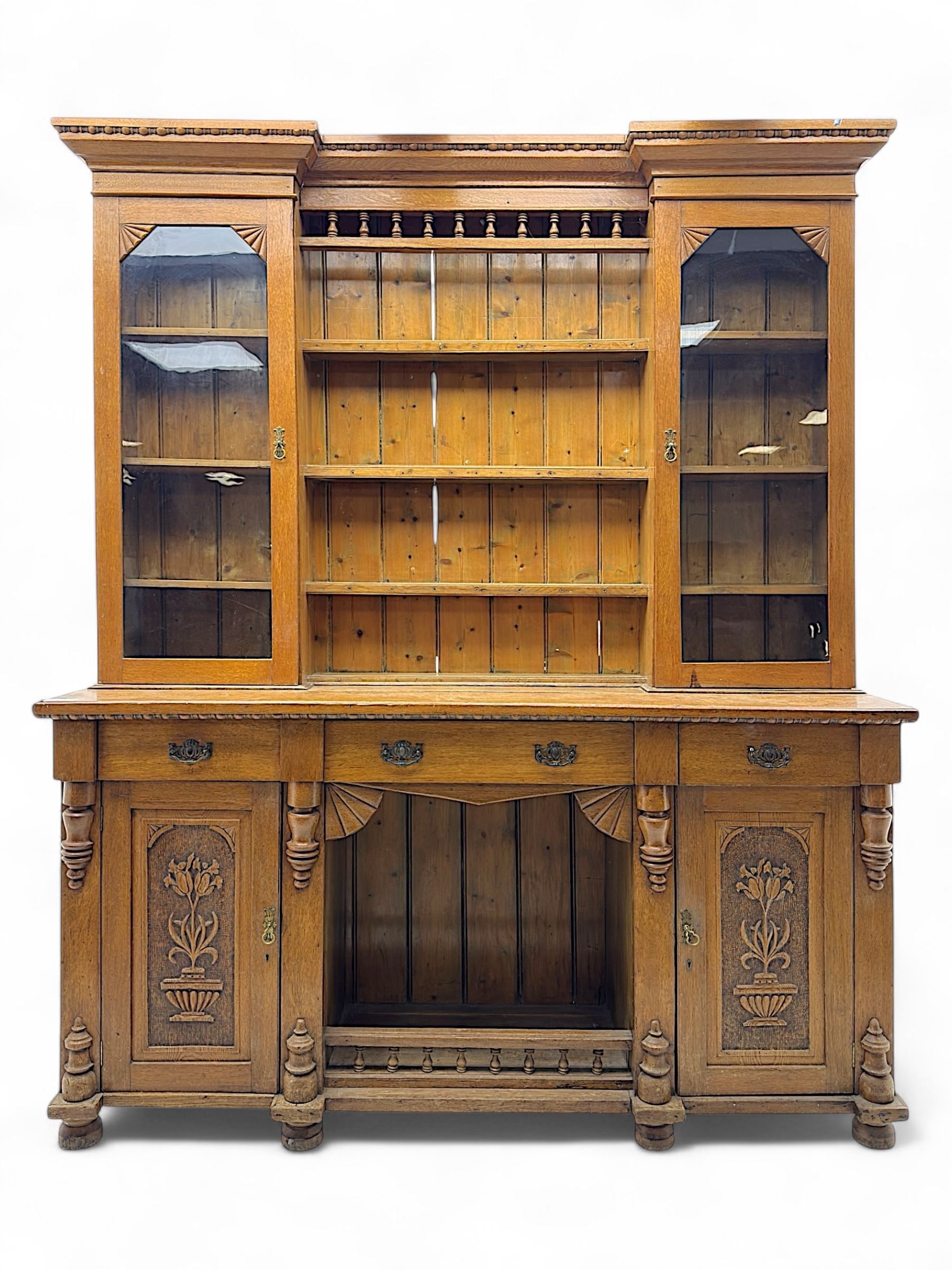 Late 19th century pine dresser, the projecting cornice with dentil and spindle moulded frieze above a three-tier plate shelves, flanked by glazed doors, the base fitted with three frieze drawers over arched apron and carved floral panelled cupboard doors, raised on turned feet