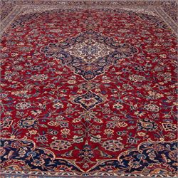 Central Persian Kashan hand-knotted carpet, madder red field with central cusped medallion in navy and ivory enclosing a floral rosette and radiating scrolling arabesques and palmettes, across a densely ornamented ground of finely drawn vinery enriched with a profusion of flowering sprays and stylized blossoms, spandrels in deep indigo and principal border of navy ground decorated with a continuous palmette and vine-scroll flanked by multiple guard stripes, with cotton fringe ends