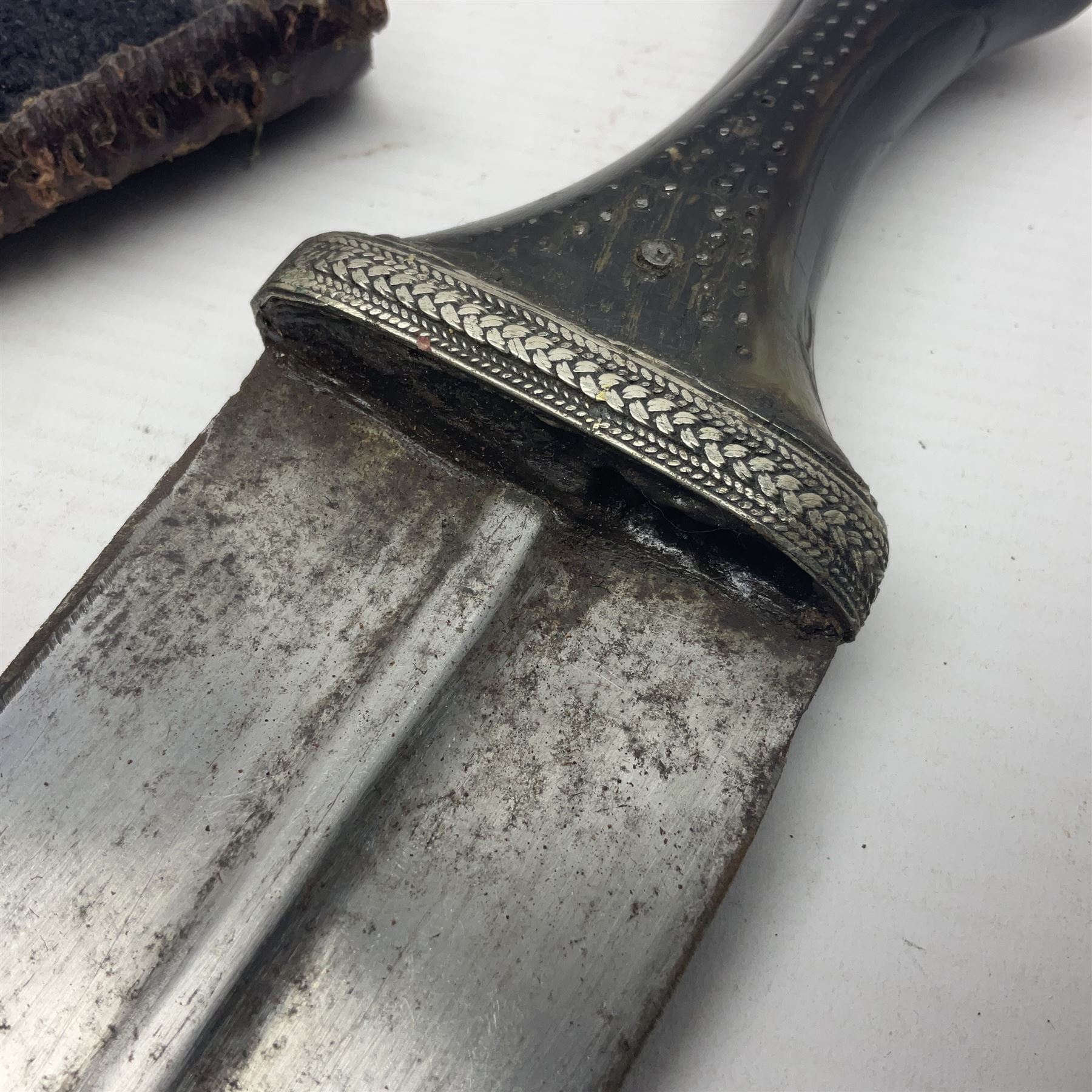 Eastern Jambiya, the 19cm double edge steel blade with raised medial ridge, waisted horn grip with pique work and white metal band; in stitched leather scabbard L33cm overall
