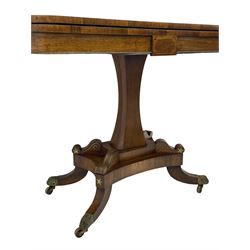 Regency rosewood and brass inlaid card table, the rectangular fold-over and swivelling top with rounded corners with a satinwood band, opening to reveal a blue baize lined interior playing surface, the frieze with a raised panel with satinwood stringing and a lower beaded brass edge, the concave and canted column with a shaped platform base applied with scrolls with brass star inlays and stringing, quadruple splayed supports terminating in foliate cast cups and castors