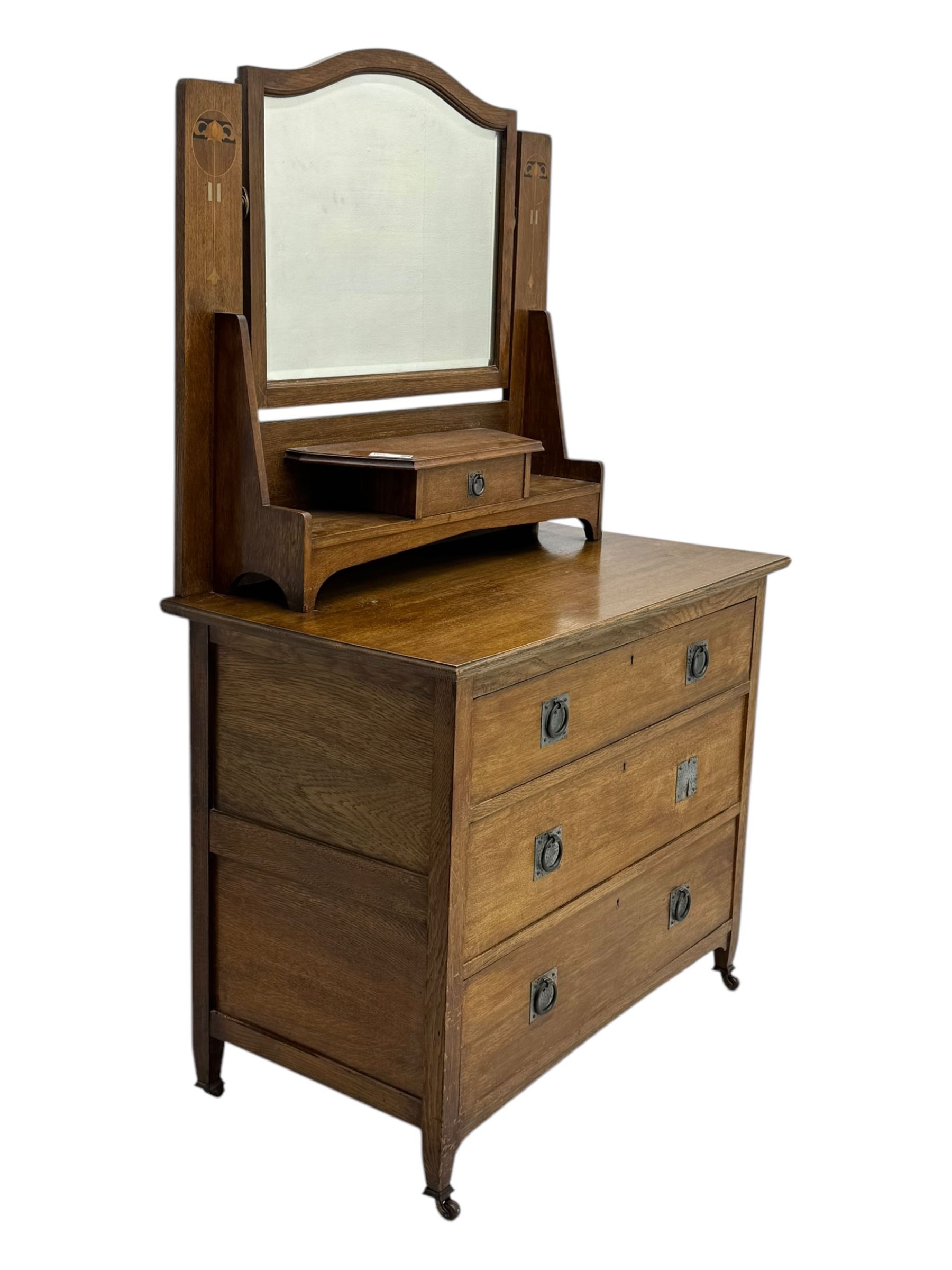 Early 20th century Arts and Crafts oak dressing chest, the arched mirror flanked by floral marquetry inlaid uprights above a shelf and drawer, over three long drawers with square hammered metal plates and circular pulls, on slender supports with castors