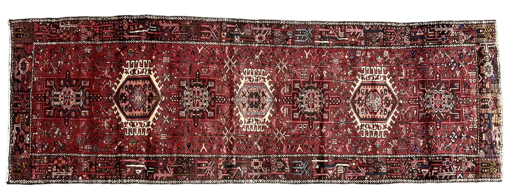 Persian Karajeh crimson ground runner rug, the field set with seven geometric lozenge medallions, the field decorated all over with stylised plant motifs, the banded border with repeating geometric shapes and patterns