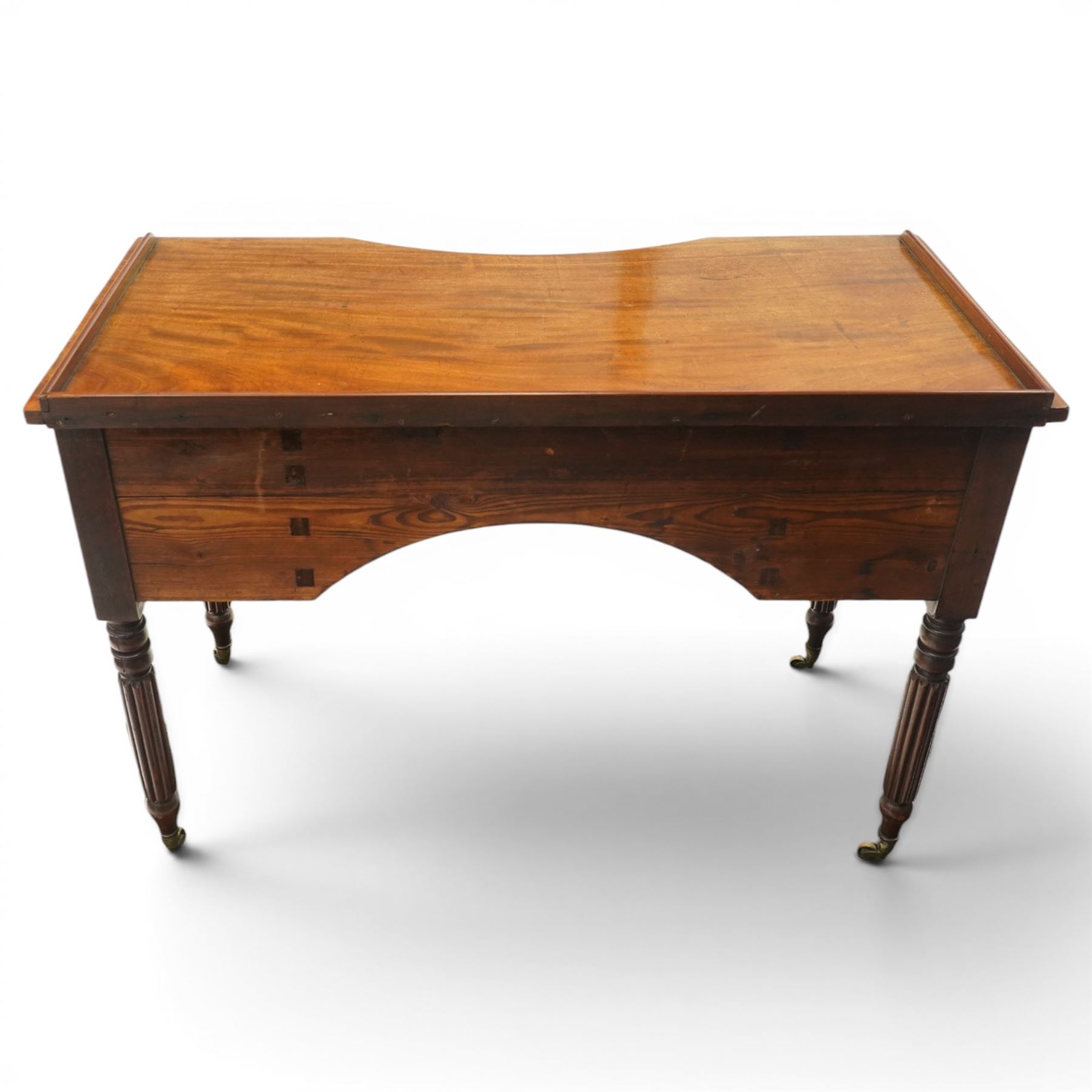 In the manner of Gillows - Regency period mahogany kneehole dressing table or writing desk, the shaped rectangular top with raised three-quarter gallery, fitted with central concave frieze drawer flaked by two graduating drawers to each side, each with ebony stringing, raised on turned and reeded supports terminating to brass cups and castors