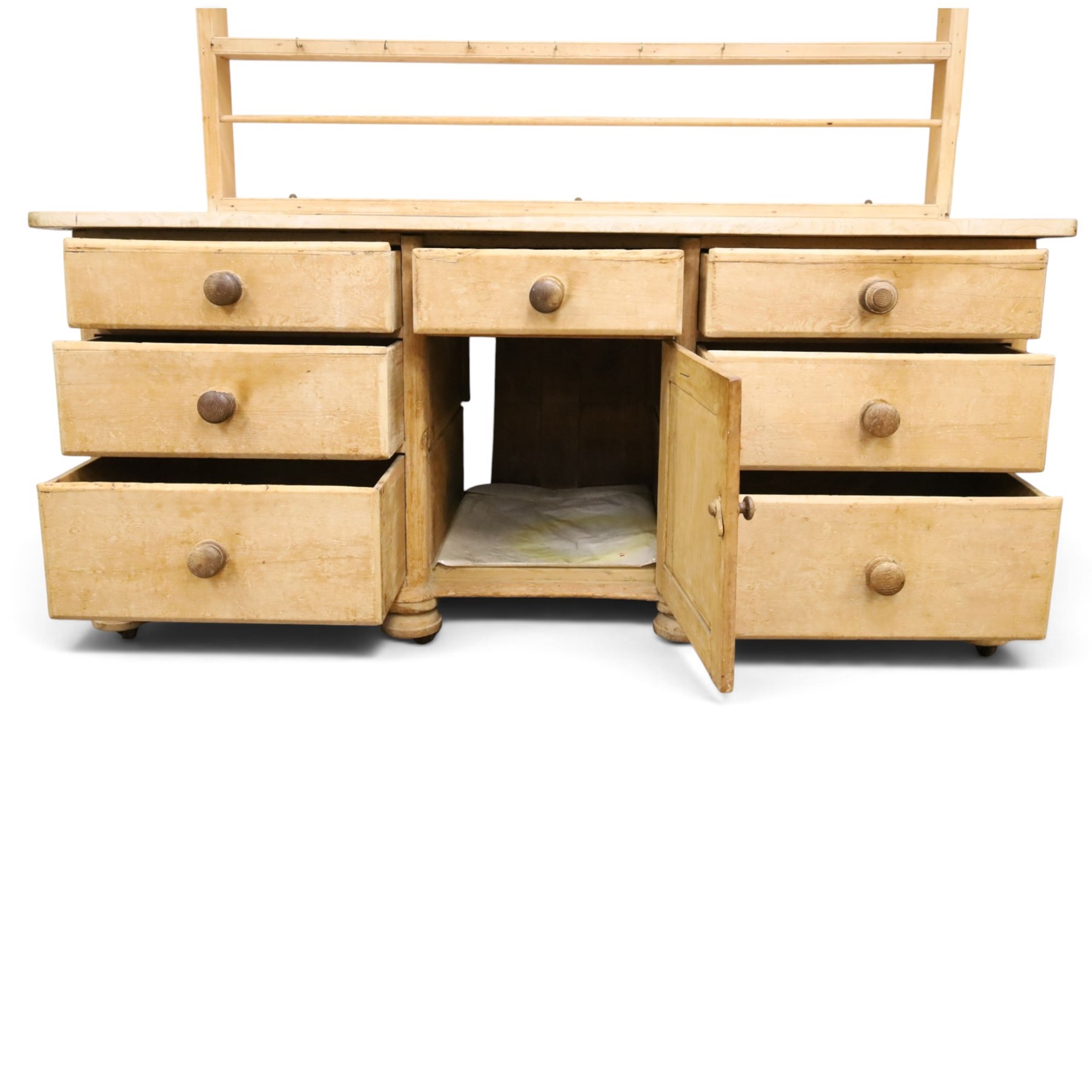 Victorian stripped pine dresser base with plate rack, the four-tier plate rack with supporting rails and hooks, the inverted-breakfront base fitted with seven assorted graduating drawers surrounding a recessed cupboard, on compressed bun feet with castors