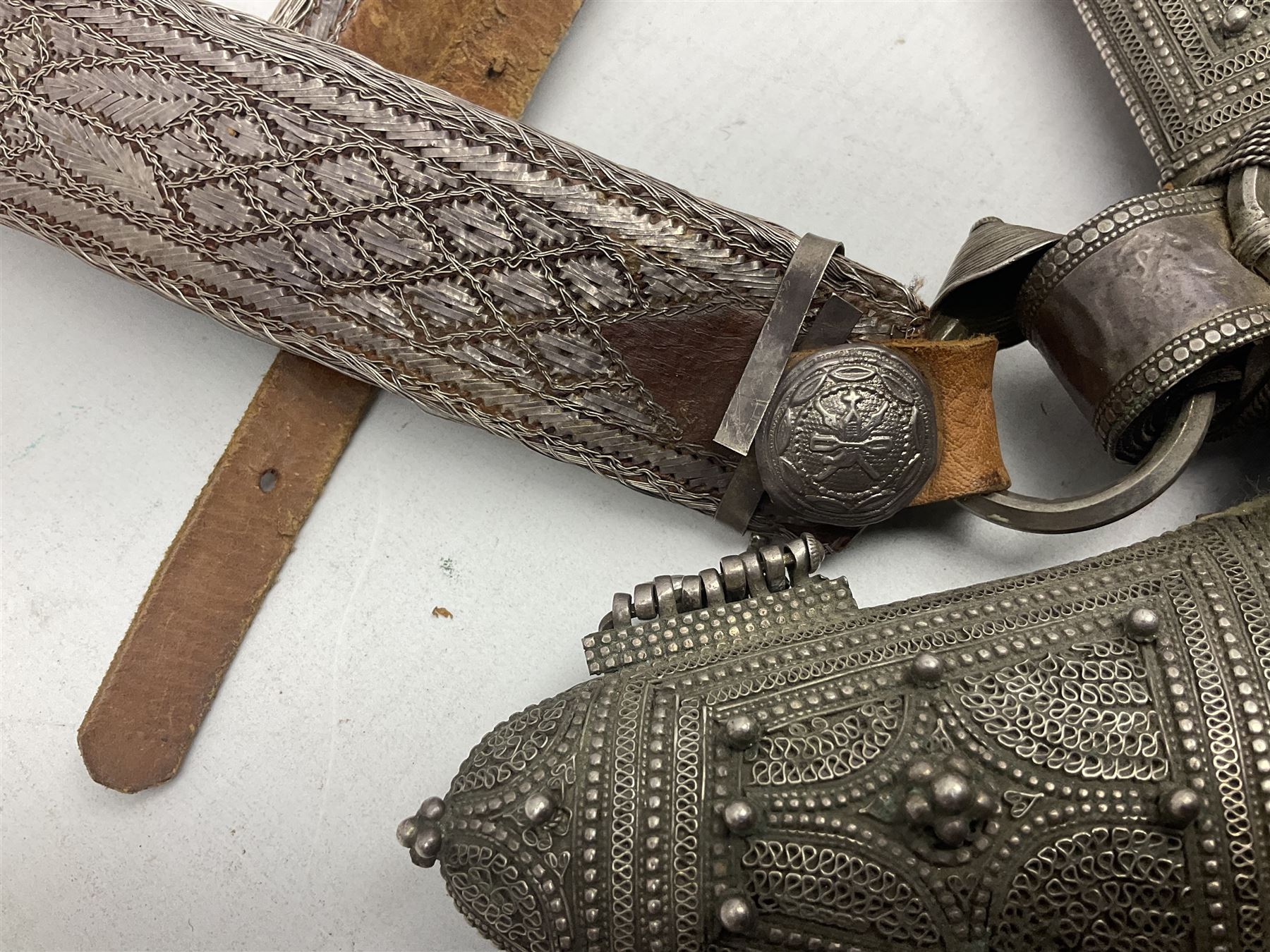 Omani Khanjar dagger, the 19cm curved blade with pronounced medial rib to either side, waisted hilt with dome shaped pommel and silver covered horn grip decorated with filigree bands, floral motifs and beadwork; in an intricately filigree decorated silver and leather scabbard with silver rings woven into the belt mount and fitted with original silver thread-work belt L34cm overall belt length 94cm