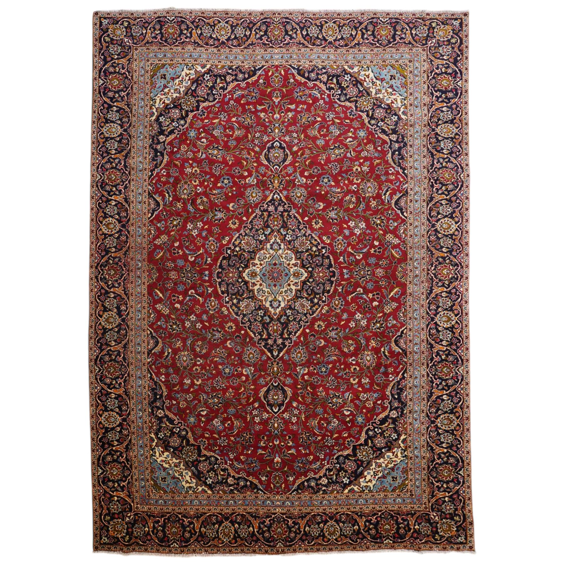 Central Persian Kashan hand-knotted carpet, crimson ground field, with central cusped lozenge medallion, enclosing a palmette and a floral rosette arrangement, surrounding field densely populated with scrolling arabesques, flowering vinery and stylized boteh motifs in tones of indigo, pale blue, ivory and ochre and spandrels of confirming design, flanked by multiple guard stripes, with cotton fringe ends 