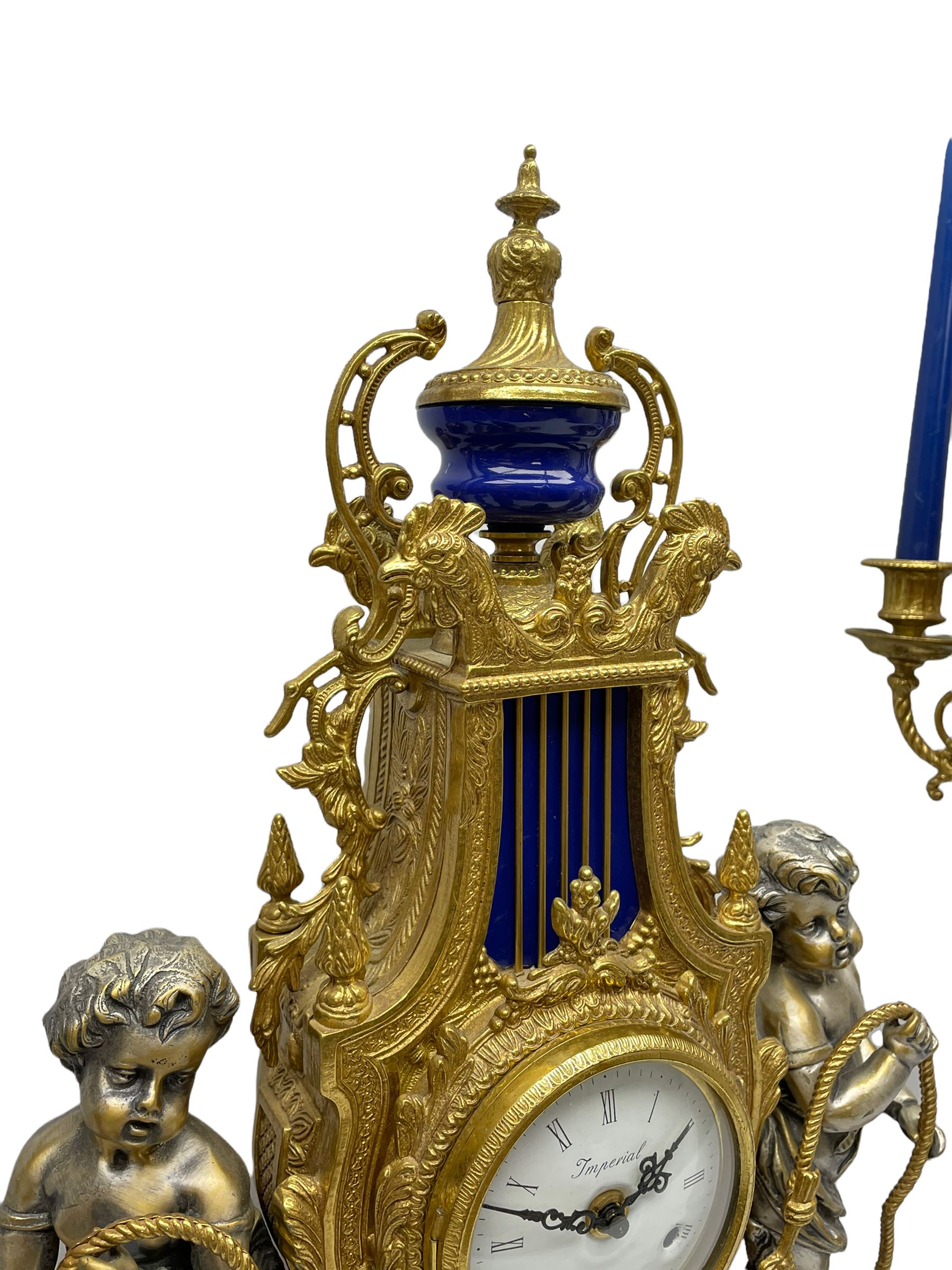 A 20th century continental gilt metal Lyre mantle clock with a pair of matching six light candelabra, spring driven movement housed in a gilt drum case on a brass and cobalt blue base with paw feet, two bronzed metal figures of mythological cherubs with animal legs and cloven hooves supporting a festooned garland swag, eight-day twin barrel striking movement with a floating lever balance escapement, striking the hours and half-hours on two bells, white enamel dial with roman numerals, minute track and pierced steel hands, dial inscribed “Imperial”. 