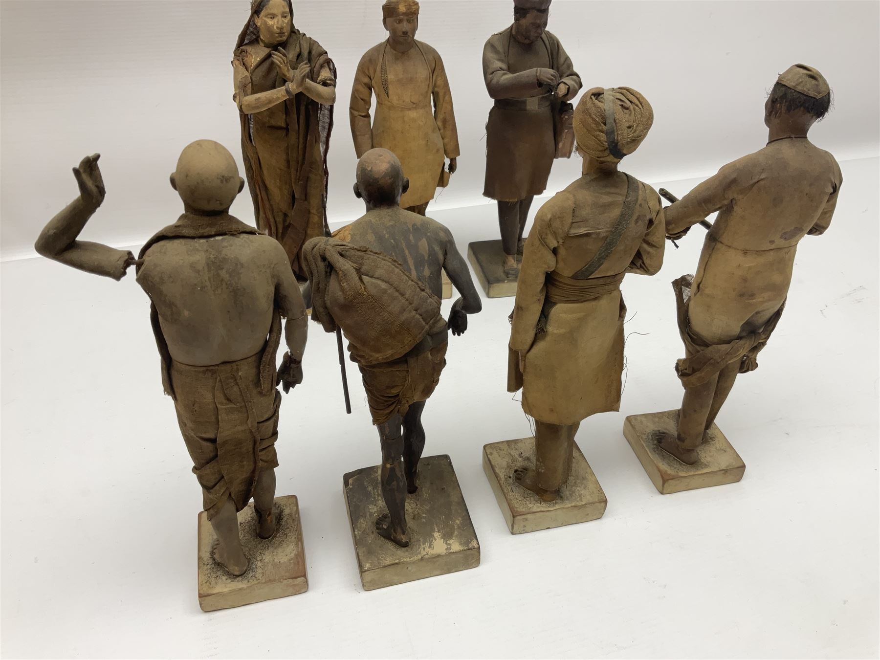 Seven 19th Century Indian clay dolls, possibly Krishnanagar, each figure with painted details, wearing cloth garments, standing on rectangular bases, some with paper label to the base, H25cm