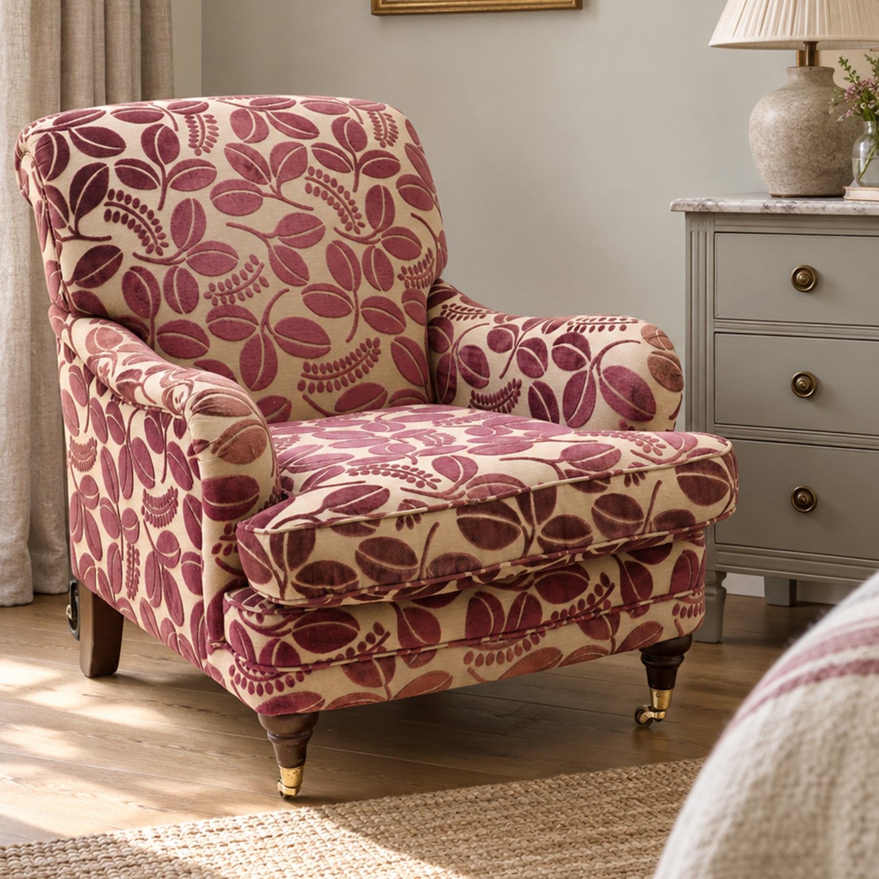 Traditional Howard shape armchair, rolled back and arms over deep seat, raised on turned supports with brass cups and castors, upholstered in plum foliate patterned fabric with loose seat cushion