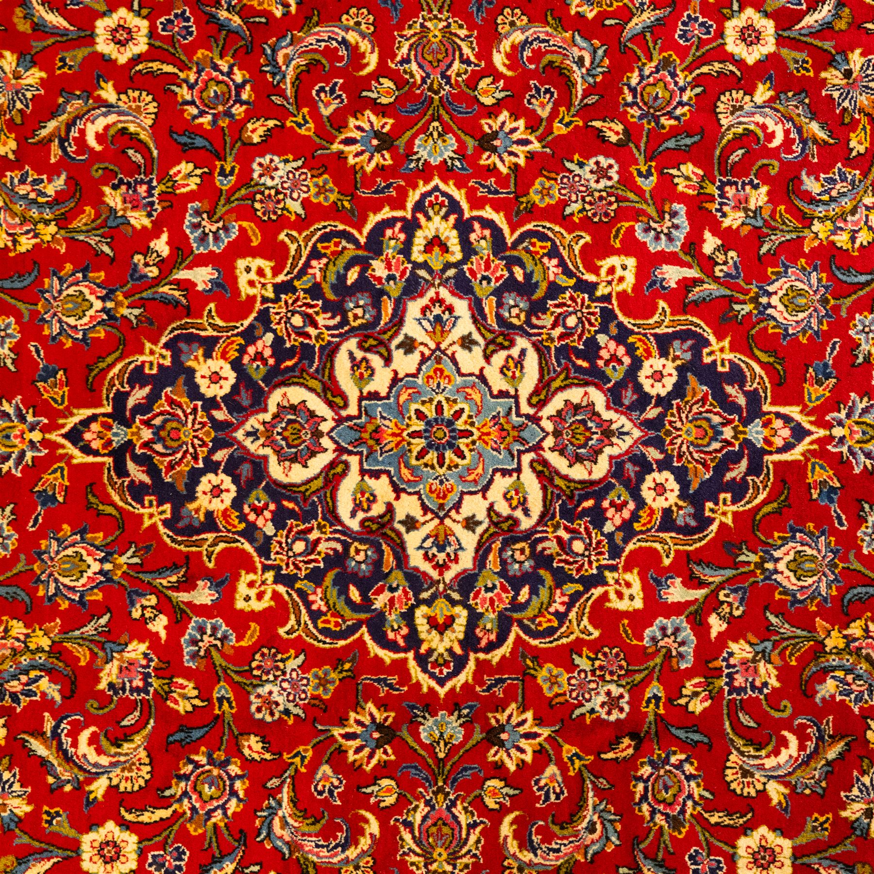 Central Persian Kashan hand-knotted carpet, central field in a ruby red ground with an indigo medallion with pendant motifs, enclosing scrolling arabesques, palmettes, and floral sprays in shades of indigo, turquoise, ivory, and gold, framed by indigo spandrels and a broad floral border between multiple guard stripes 