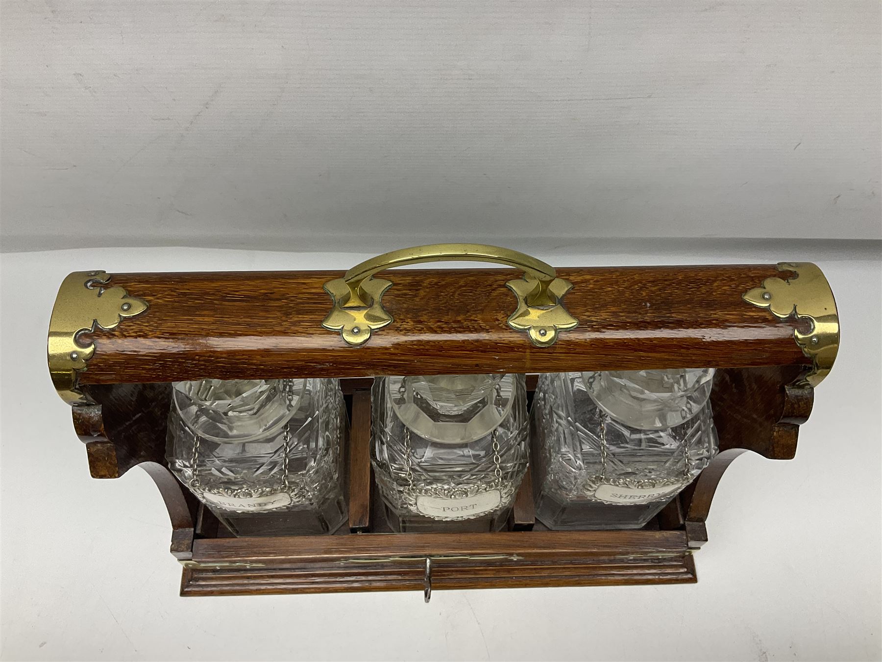 Edwardian oak three bottle tantalus, with brass mounts and three square sided cut glass decanters each with hallmarked silver decanter labels, complete with key, H31cm