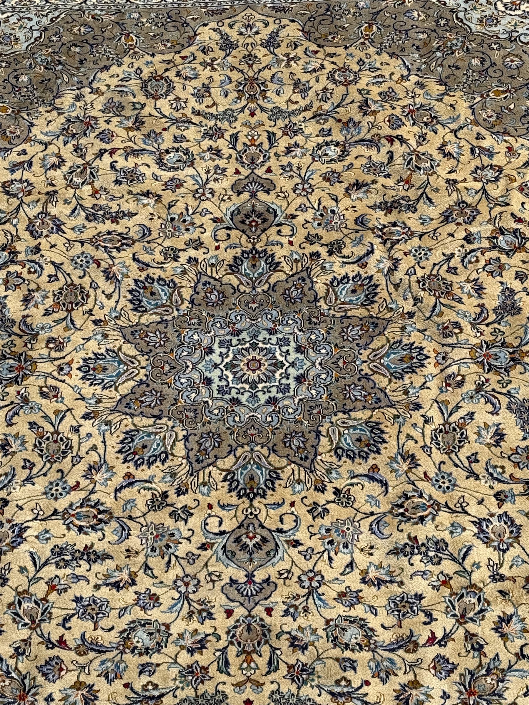 Central Persian Kashan pale khaki and ivory ground carpet, shaped central medallion enclosing small stylised motifs, the field decorated profusely with trailing foliate branches and plant motifs, guarded border with repeating floral design