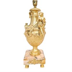 Pair of Rococo style gilt metal table lamps, the urn shaped bodies each with applied figure seated upon shoulder, and female mask to opposite side issuing scroll leading to top of neck, the body cast and applied with stiff leaf detail, and foliate swags and wreaths, upon square pink marble plinths with applied gilt metal mask mounts, and four gilt metal bracket feet, each supported a pleated cream silk closed top lampshade, H63cm
