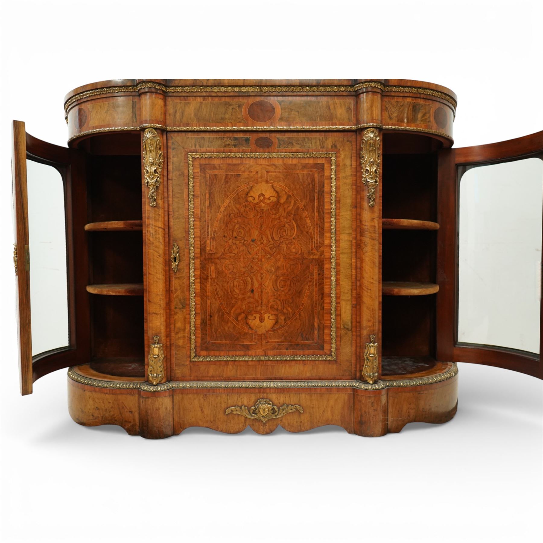 Late Victorian figured walnut credenza, the shaped demi-lune top with cross-banding and stringing over an inlaid frieze with foliate gilt-metal mounts, fitted with a central geometric-inlaid panelled door, flanked by convex-glazed side-cabinets with a gilt metal slip and outer-banding, the uprights surmounted by gilt-metal cartouche mounts with cascading foliage details, raised on a shaped skirted base with central female-mask gilt mount