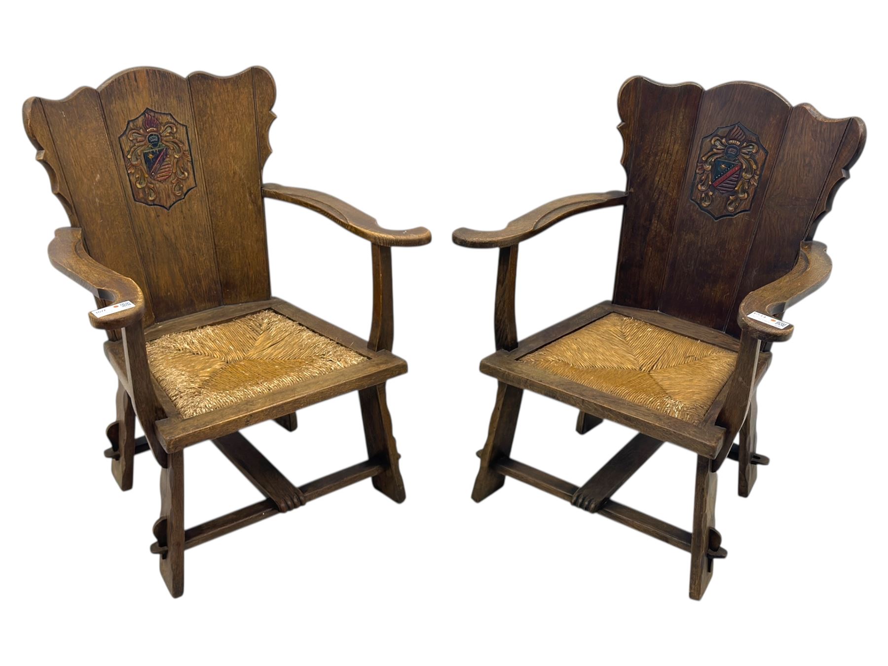 Pair of Arts and Crafts oak armchairs each with shaped backrest centred by polychrome painted crest, rush seat over straight square legs united by shaped central stretcher