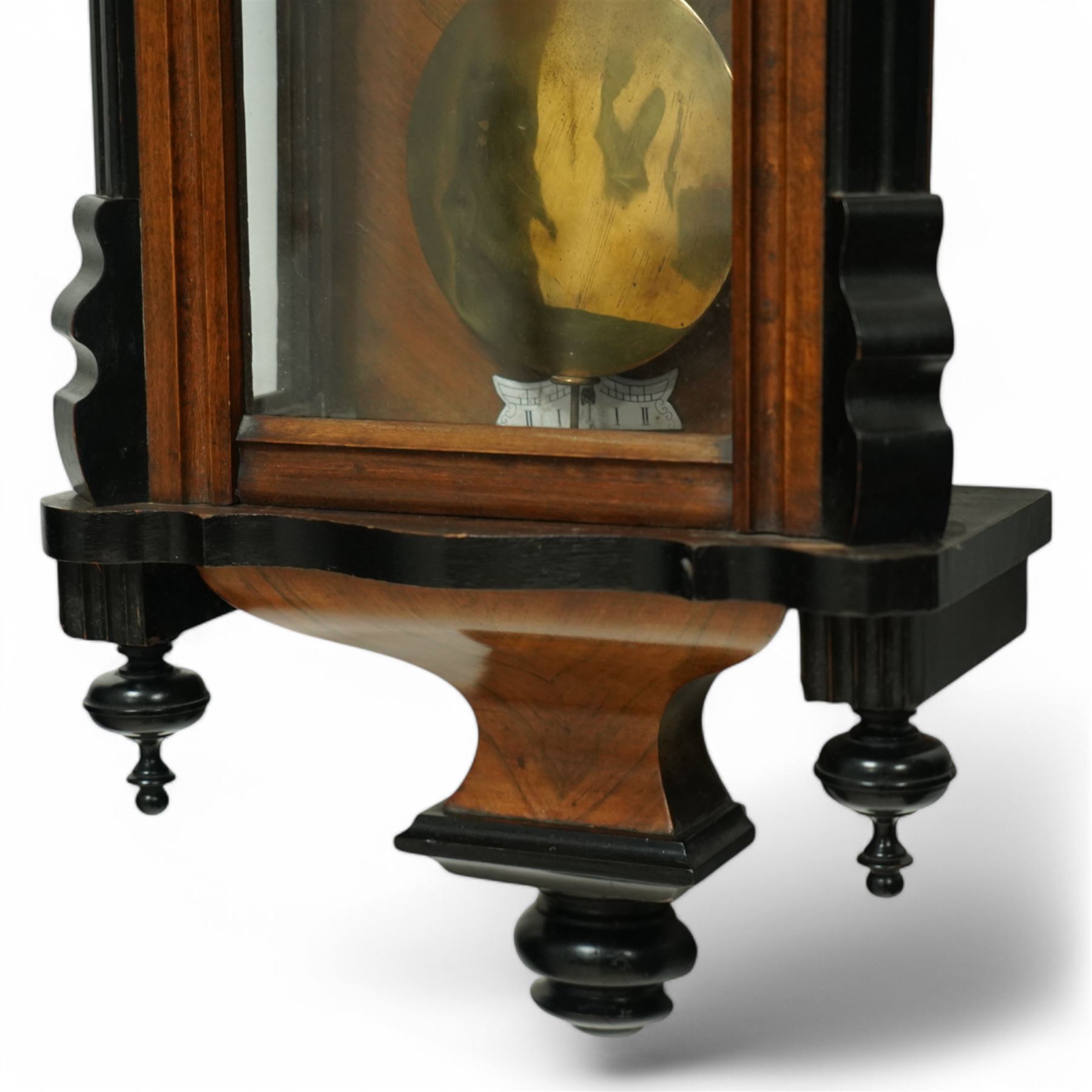 German- Late 19th century twin weight 8-day Vienna wall clock, in a walnut and ebonised case with turned finials and pendants, fully glazed door displaying the the brass cased weights and pendulum, enamel two-part dial with Roman numerals, minute rack, seconds hand and gothic steel hands, rack striking movement with a deadbeat escapement, striking the hours and half-hours on a coiled gong. With key.