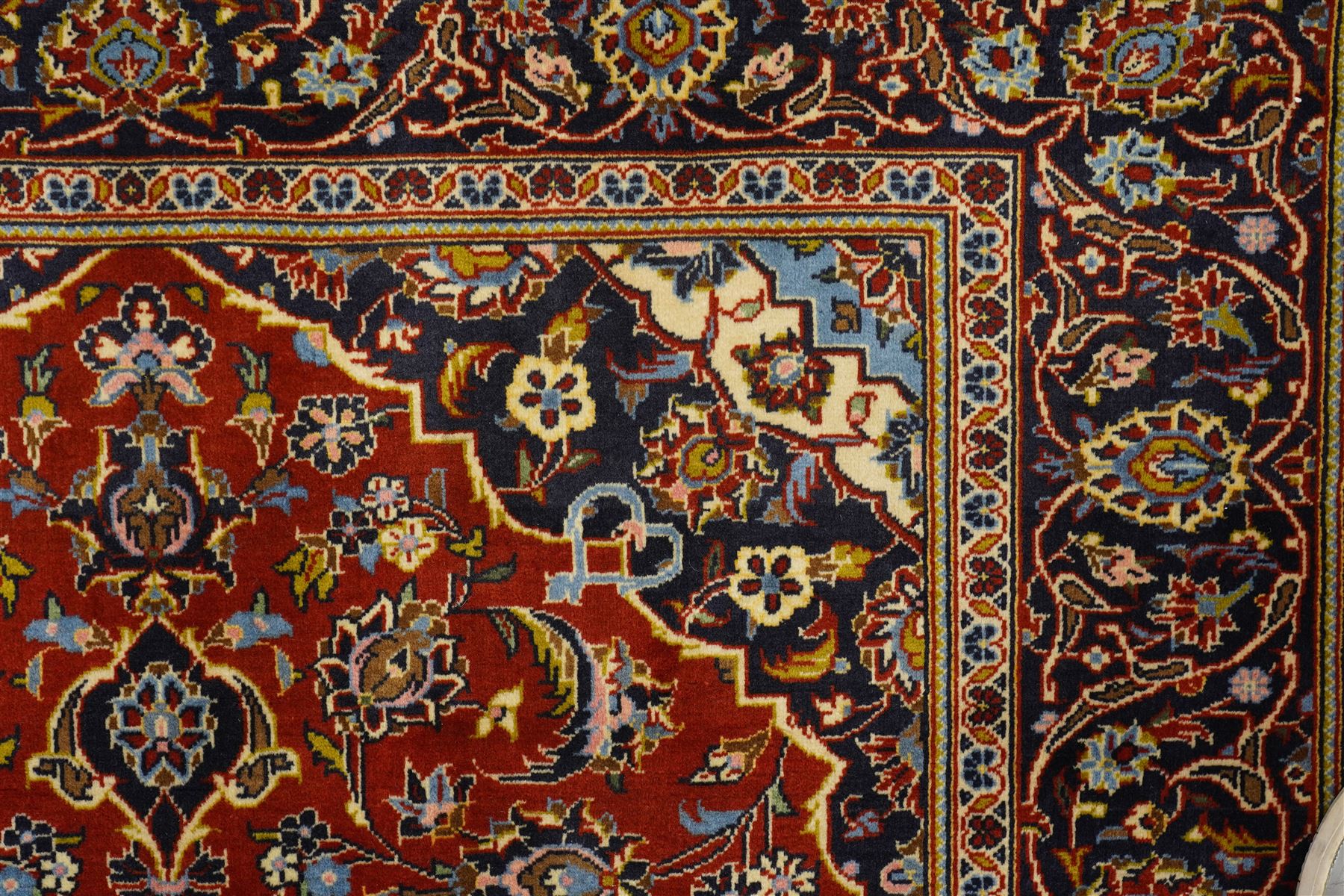 Central Persian Kashan hand-knotted rug, madder red field with central articulated cusped medallion in ivory and sky blue, issuing network of scrolling arabesques palmettes and floral sprays with conforming spandrels, indigo main border of scrolling vinery and stylized palmettes, framed by multiple guard borders, with cotton fringe ends  
