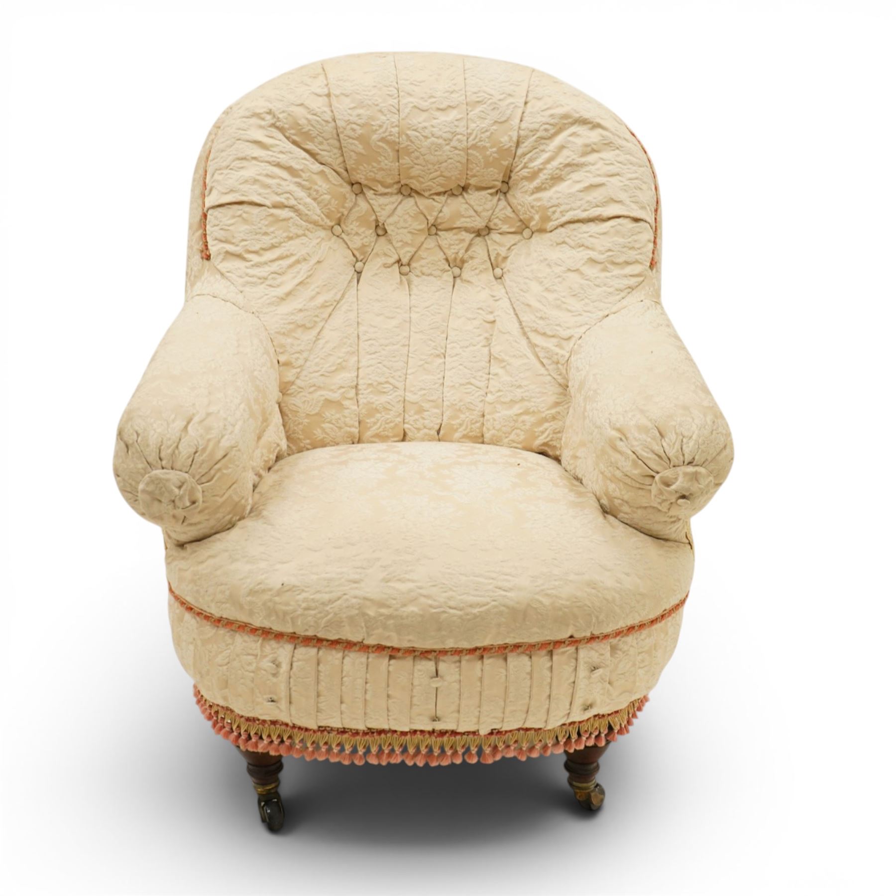 Victorian button-back nursing chair, the rounded horseshoe back enclosing a deep upholstered seat, padded arms and sprung seat in cream floral damask fabric, with crimson piping and tasselled lower fringe, on turned front legs with castors